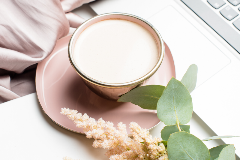 Cup of milk on pink saucer with green leaves and a flower, next to a laptop keyboard.