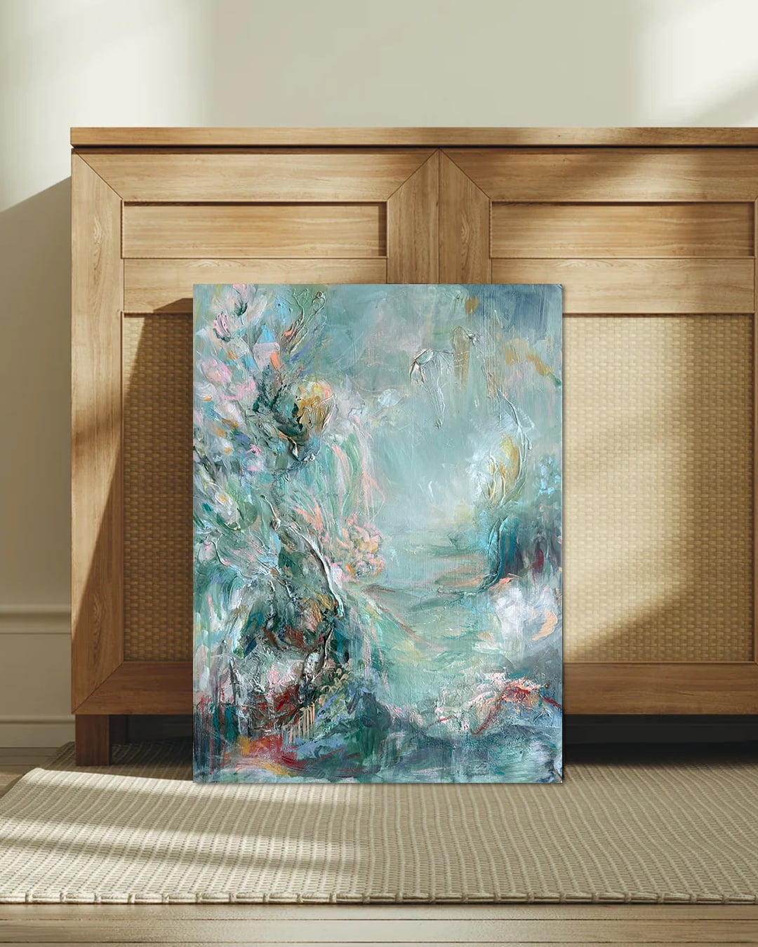 Abstract painting with blue, pink, and white colors leaning against a wooden cabinet, sunlight casting shadows on a beige rug.