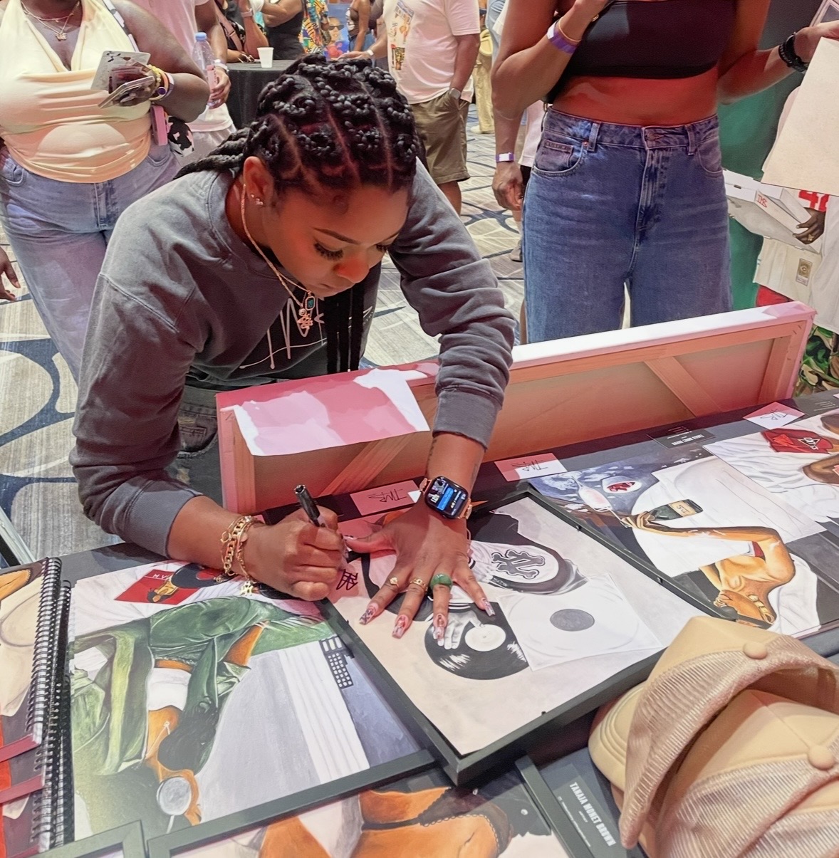 Woman signing artwork at table with people in background, wearing gray sweatshirt, with jewelry and a smartwatch.