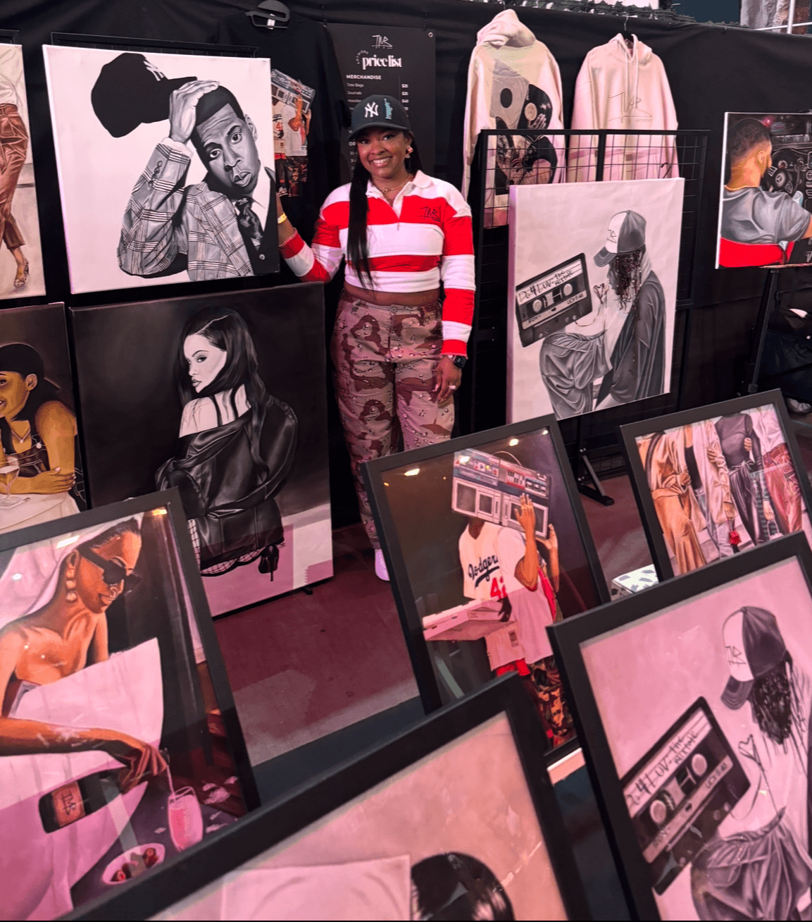 Woman standing among various framed and unframed artworks at an art exhibition, smiling, wearing a striped sweater and camouflage pants.