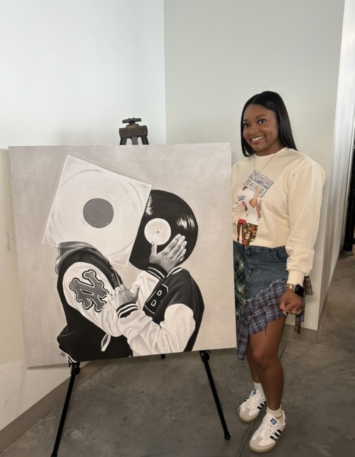 Young woman standing next to a black and white painting on an easel, smiling, in an indoor setting.