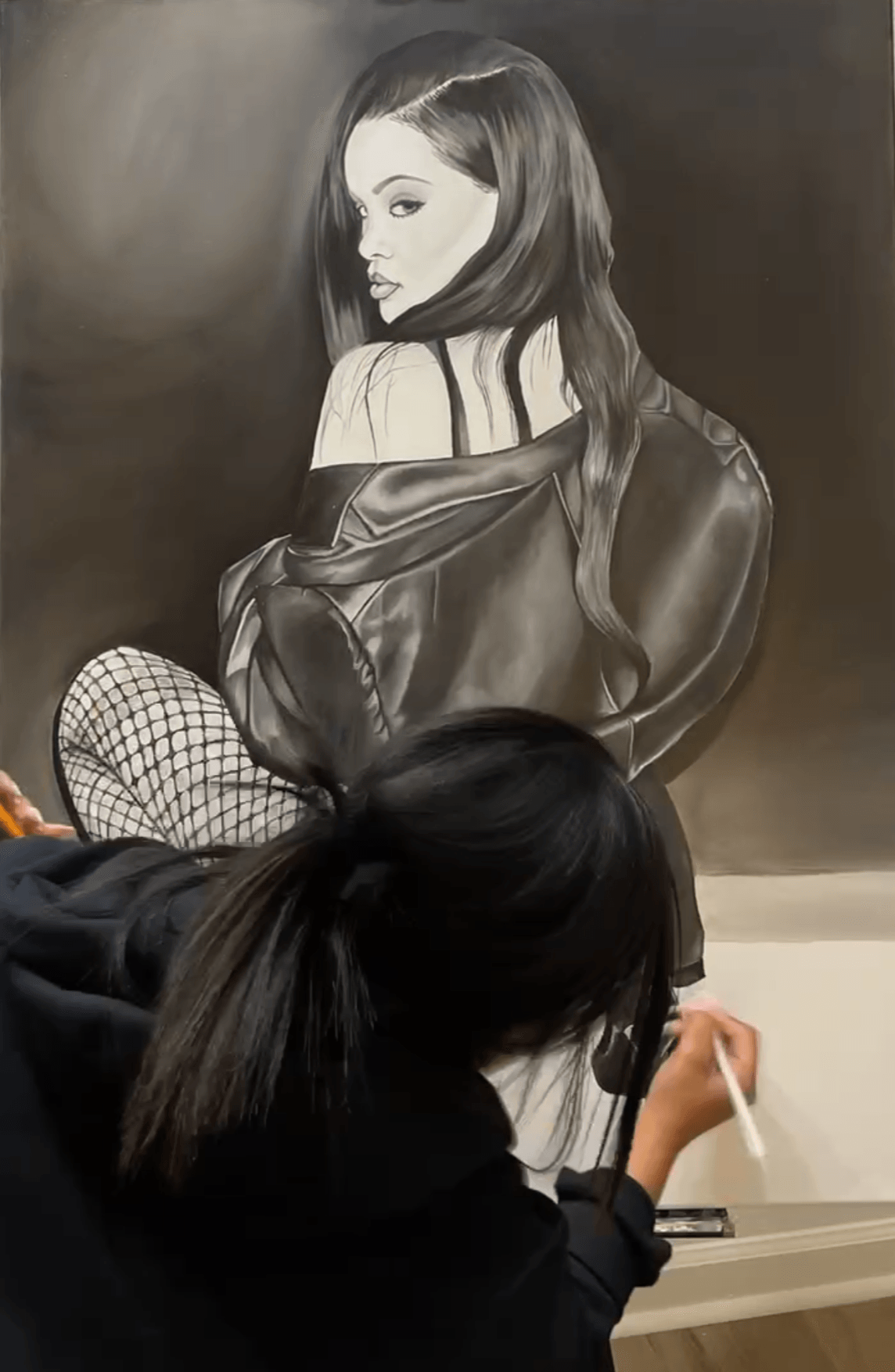 Person drawing a realistic black and white portrait of a woman with long hair, wearing an off-shoulder top, on a wall.