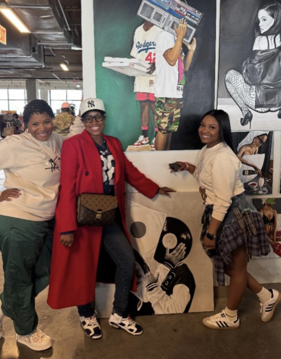 Three women standing in front of a mural with a person holding a boombox, smiling, in an indoor space.