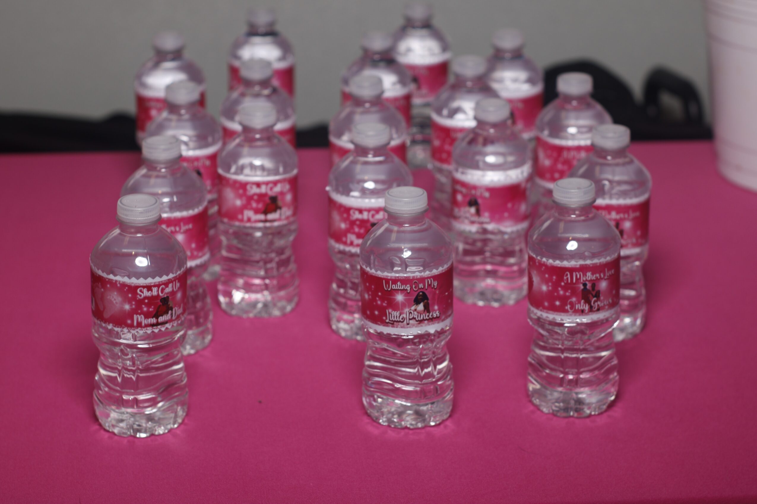 Multiple small water bottles with pink labels arranged on a pink surface, with a black background and a pink object on the right