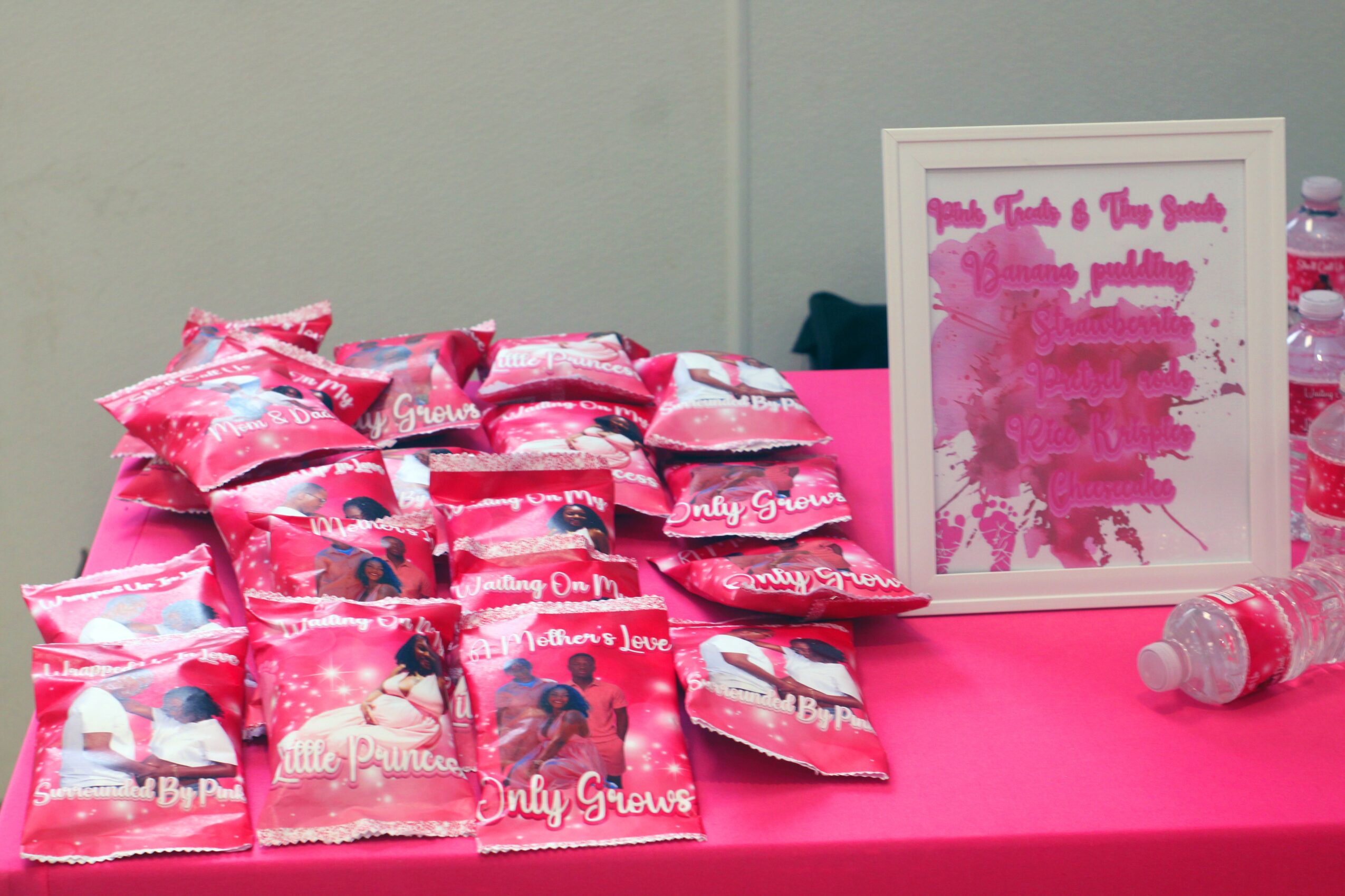 Pink-themed display with small pink bags, a framed artwork, and water bottles on a pink tablecloth.