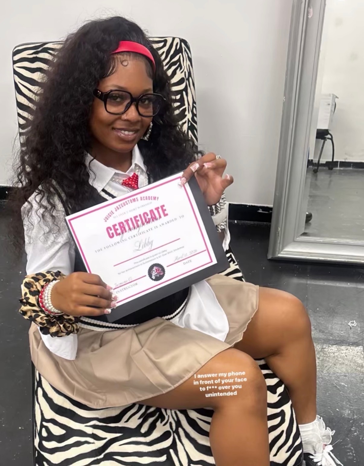 Young girl with curly hair, glasses, and a pink headband, smiling and holding a certificate, sitting on a zebra-print chair.