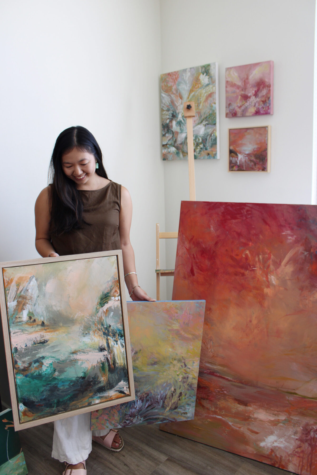 Woman with long dark hair in a brown top stands in an art gallery with colorful paintings, some leaning against the wall.