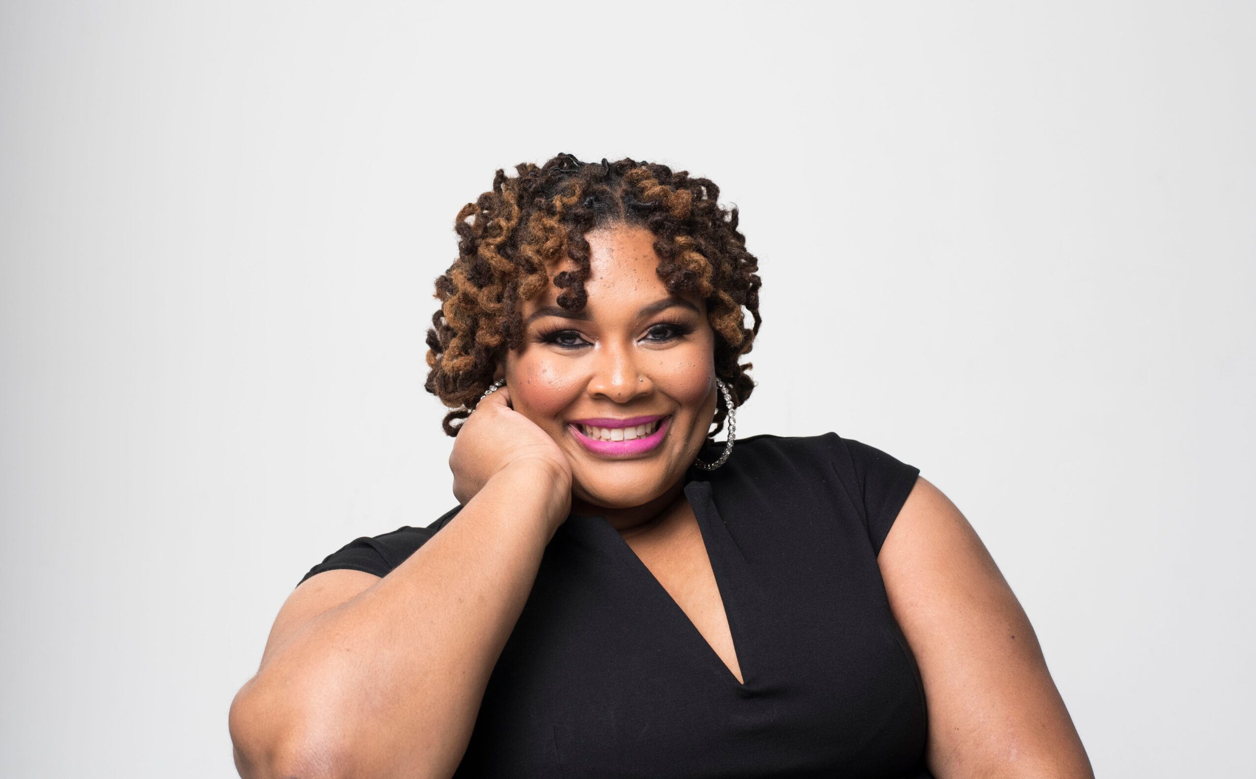 Woman with curly hair smiling, resting her head on her hand, wearing a black top, against a plain white background.