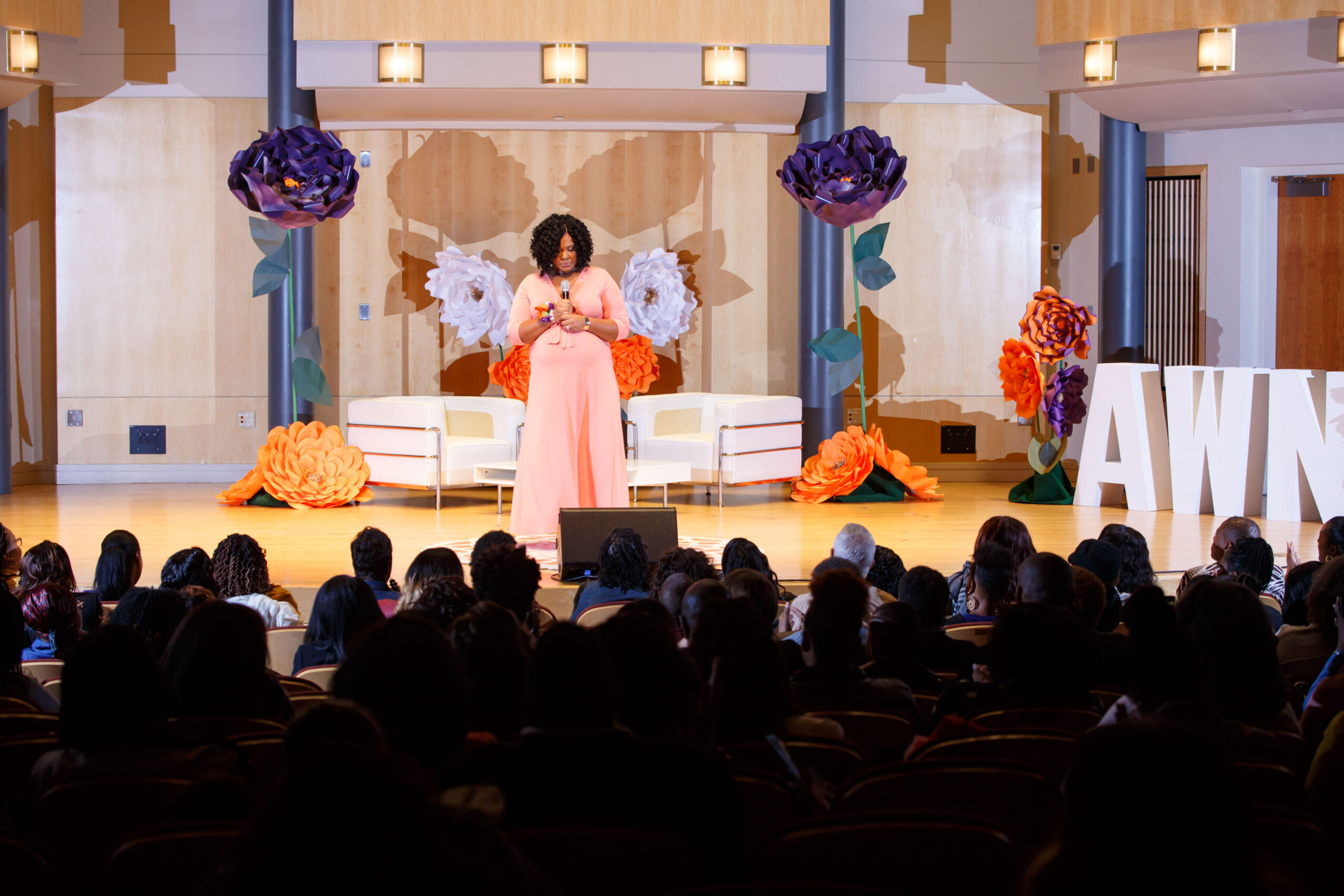A woman in a pink dress stands on stage with large floral decorations and an audience watching.