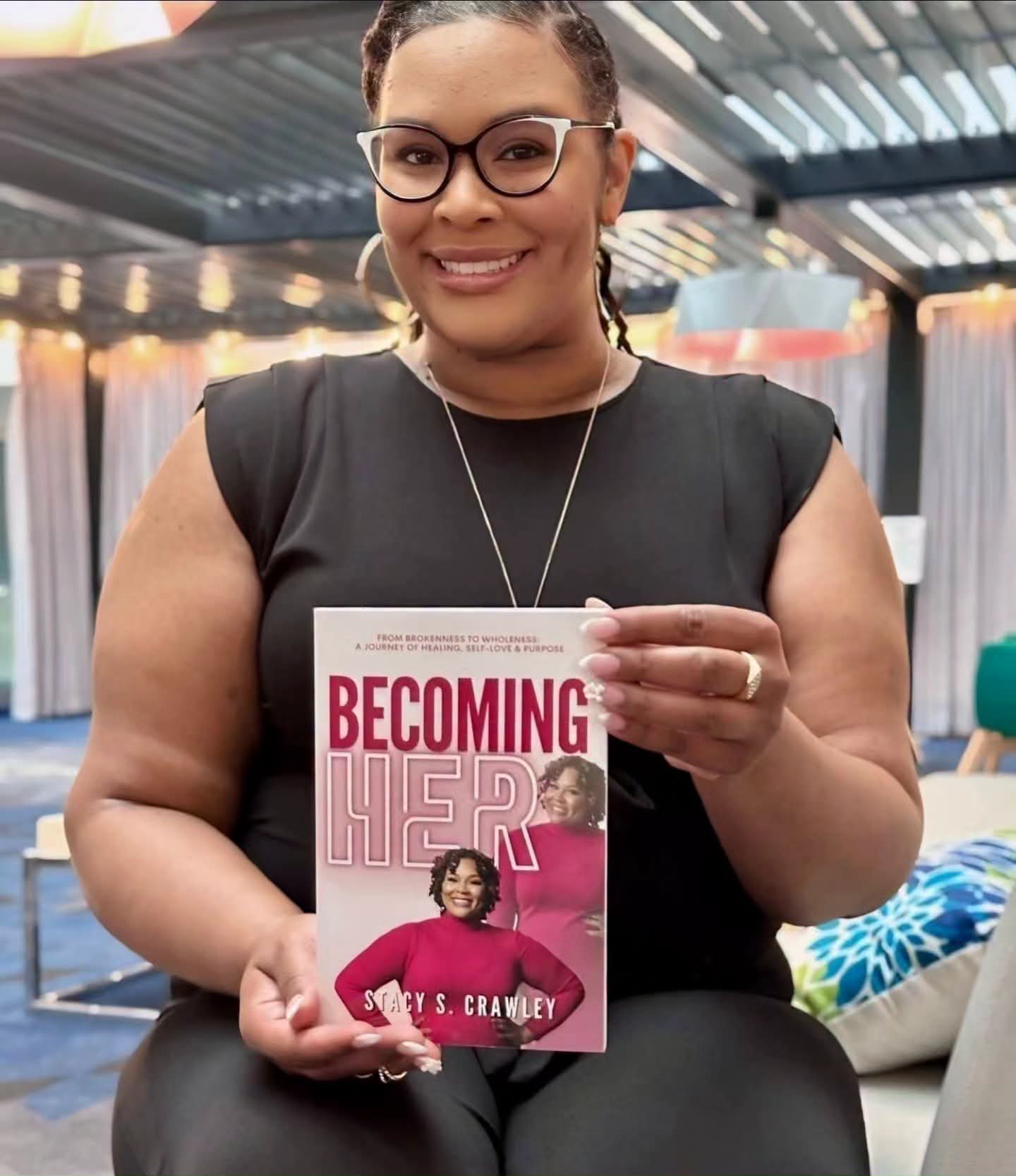 Woman with glasses holding a book titled 'Becoming Her' in a room with colorful decor.