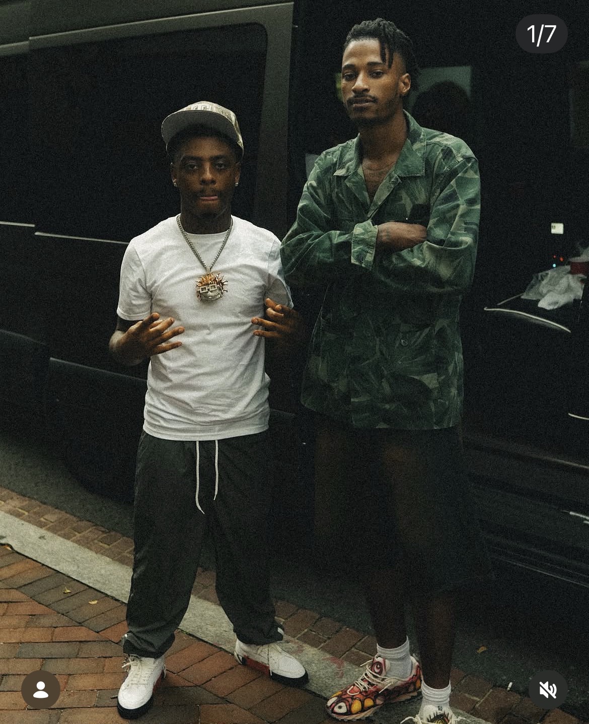 Two young men stand side by side in front of a black vehicle, one with arms crossed, the other making a hand gesture.