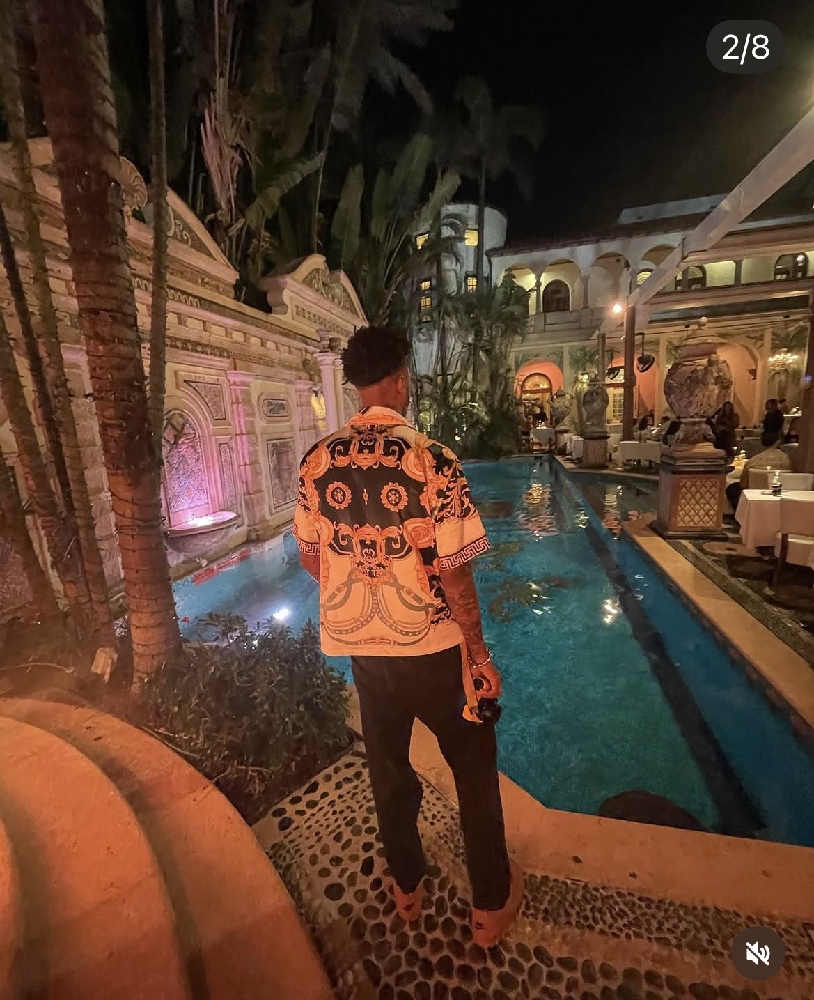 Person standing near a pool at night with decorative architecture and lush greenery in the background.
