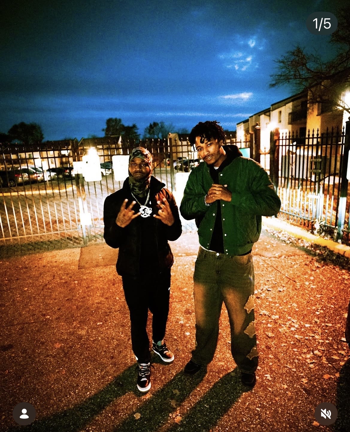 Two people standing outdoors at night, illuminated by streetlights, with a fence and buildings in the background.