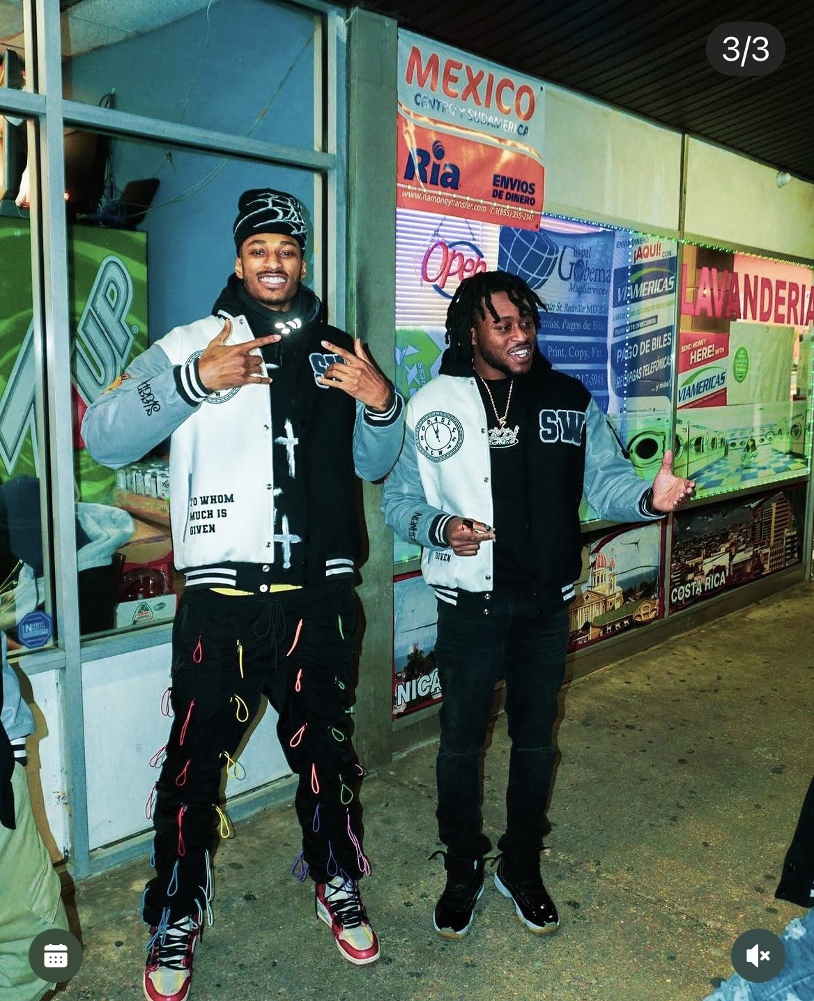 Two young men stand outside a store with colorful signs, smiling and making hand gestures. One wears a black and white jacket, the other a black and white hoodie.