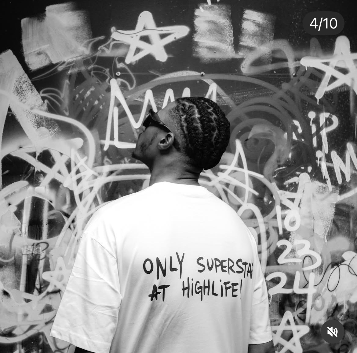 Person with braided hair wearing a T-shirt with text, standing in front of a graffiti wall.