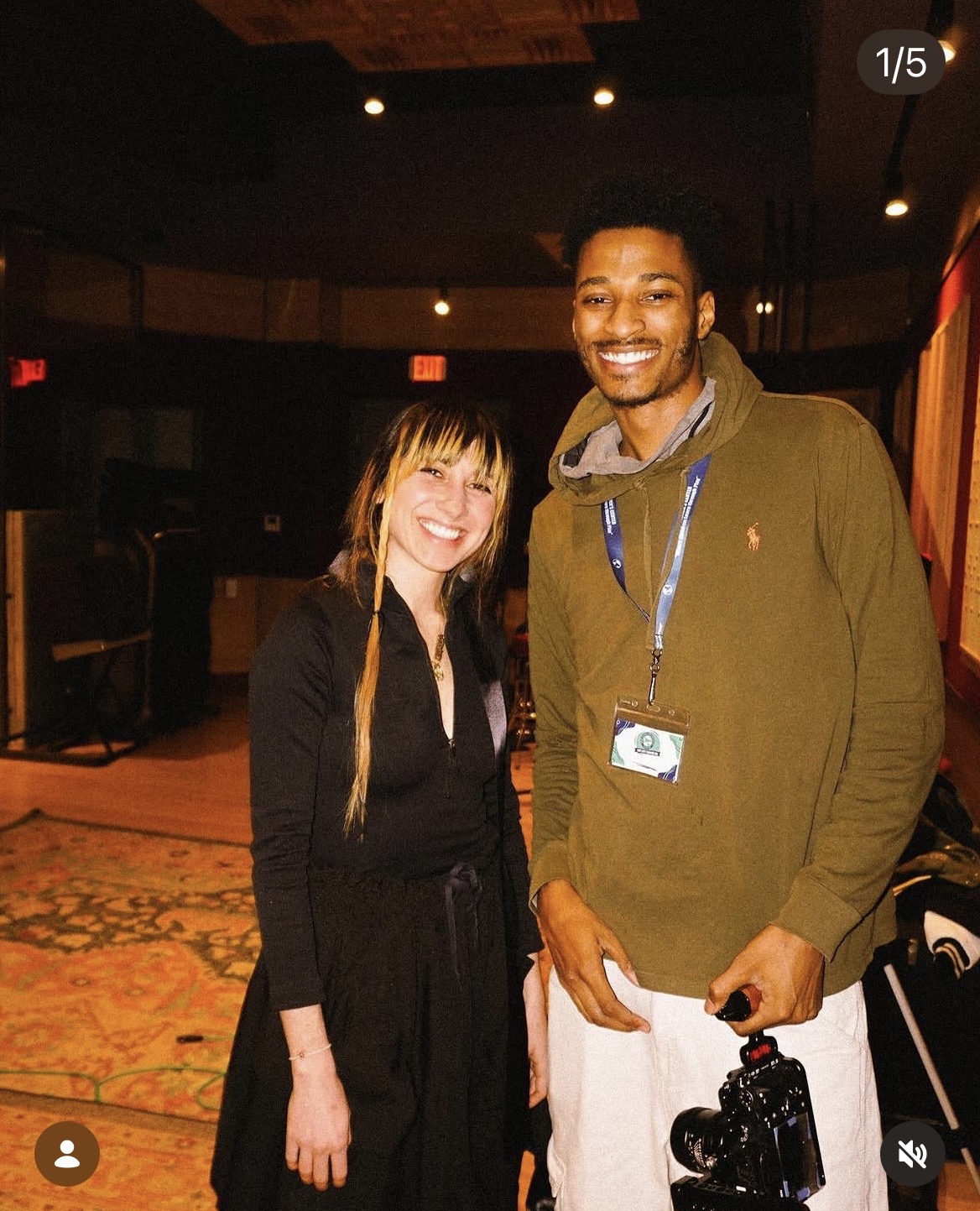 Two smiling people standing indoors, one woman with long hair and a man with a camera, both wearing casual clothing.