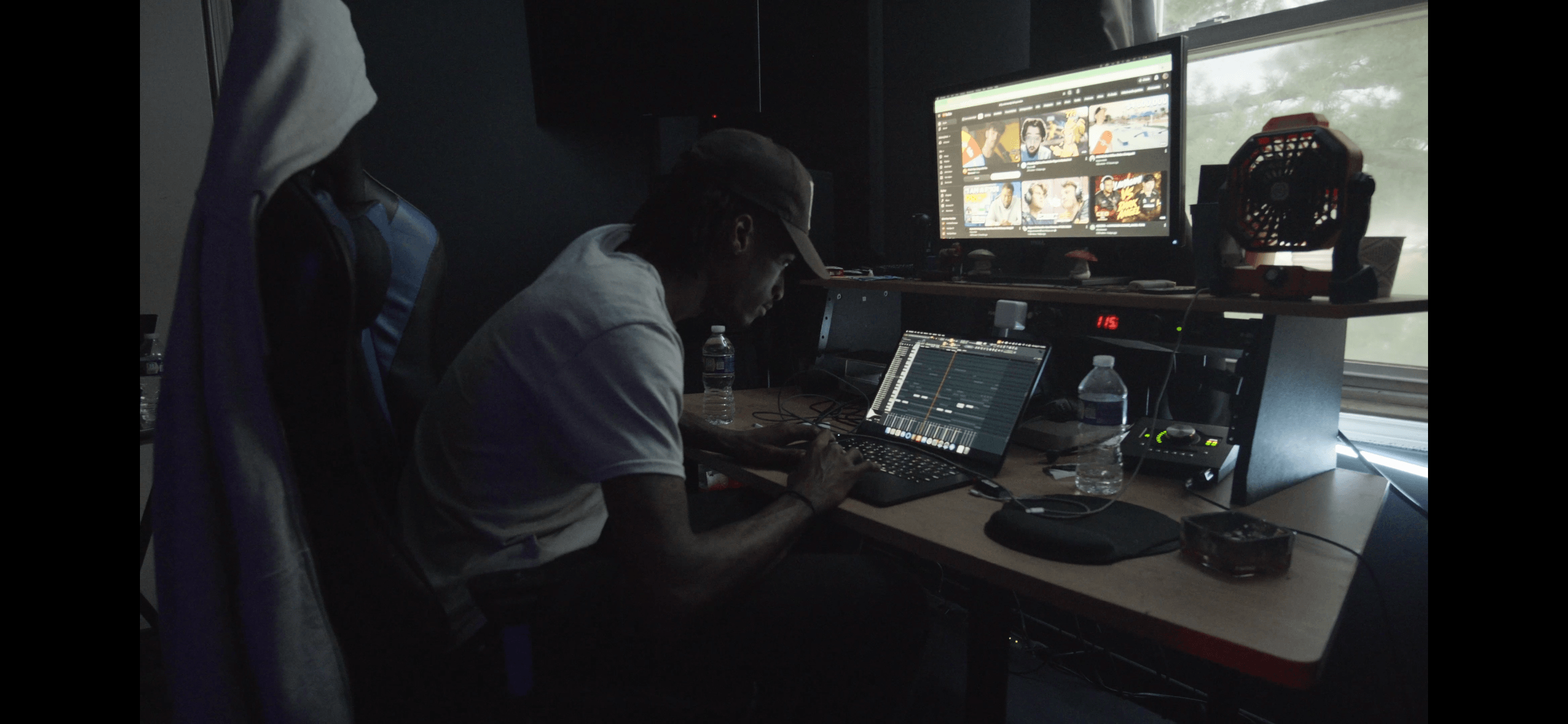 Person working at a desk with multiple monitors, editing video or images, in a dark room, with a water bottle nearby.