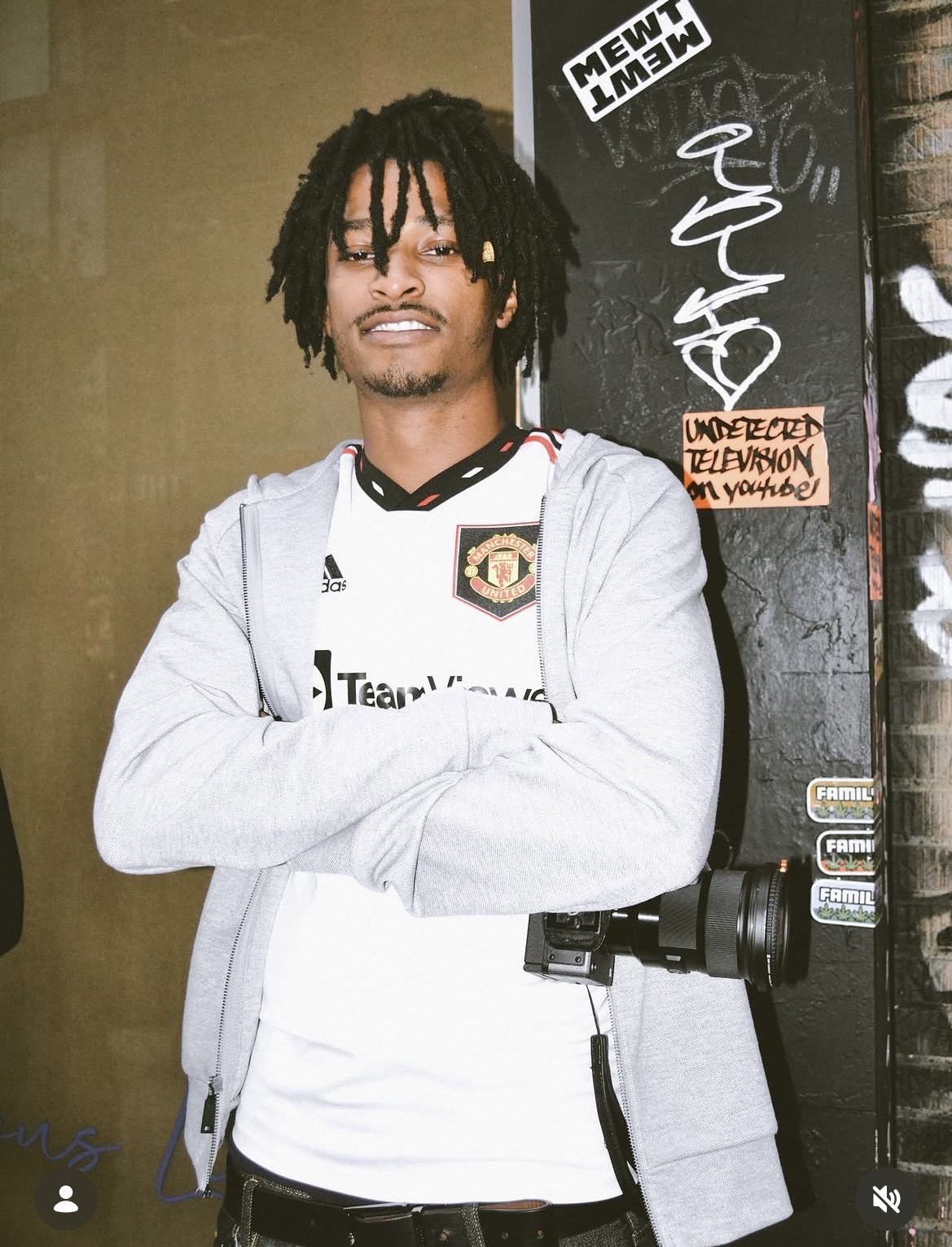 Young man with dreadlocks standing with arms crossed, wearing a Manchester United jersey and light gray hoodie, in front of a black wall with stickers.
