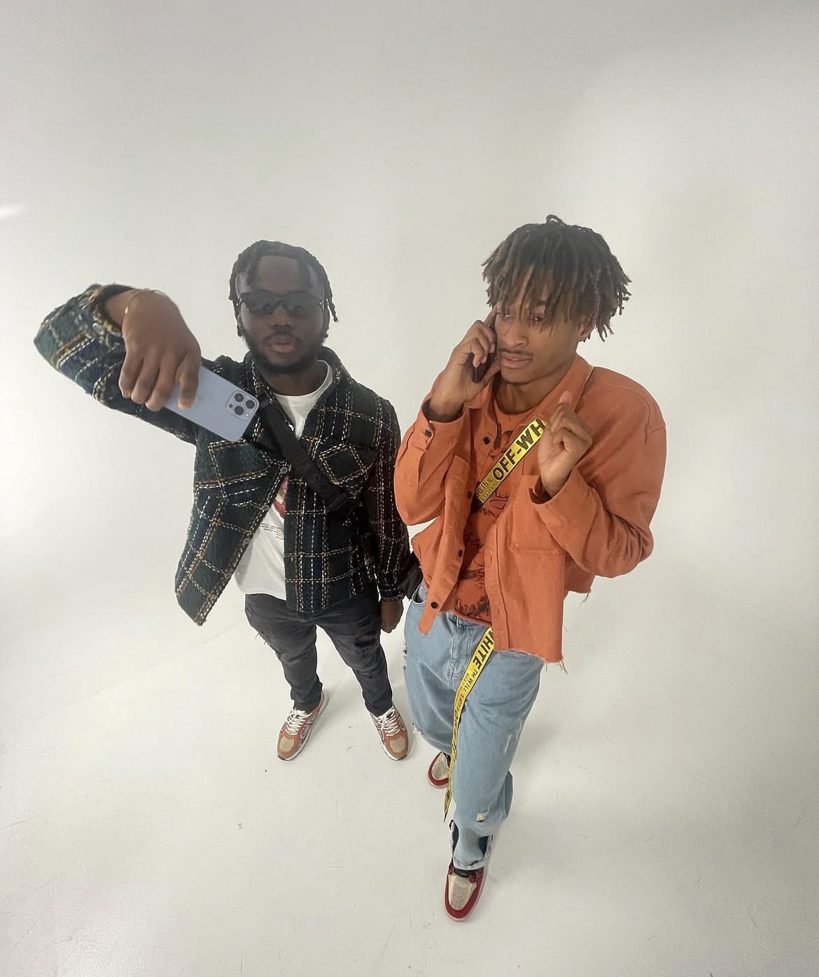 Two young men taking a selfie against a plain white background, one wearing sunglasses and a plaid shirt, the other in an orange jacket.