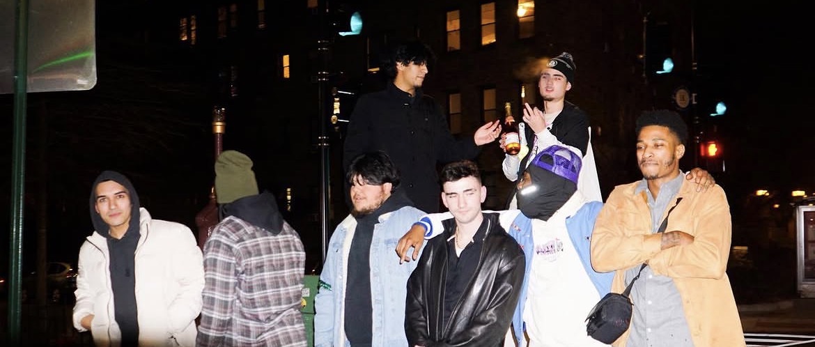 Group of seven young people standing outdoors at night, some smiling and talking, city lights in background.
