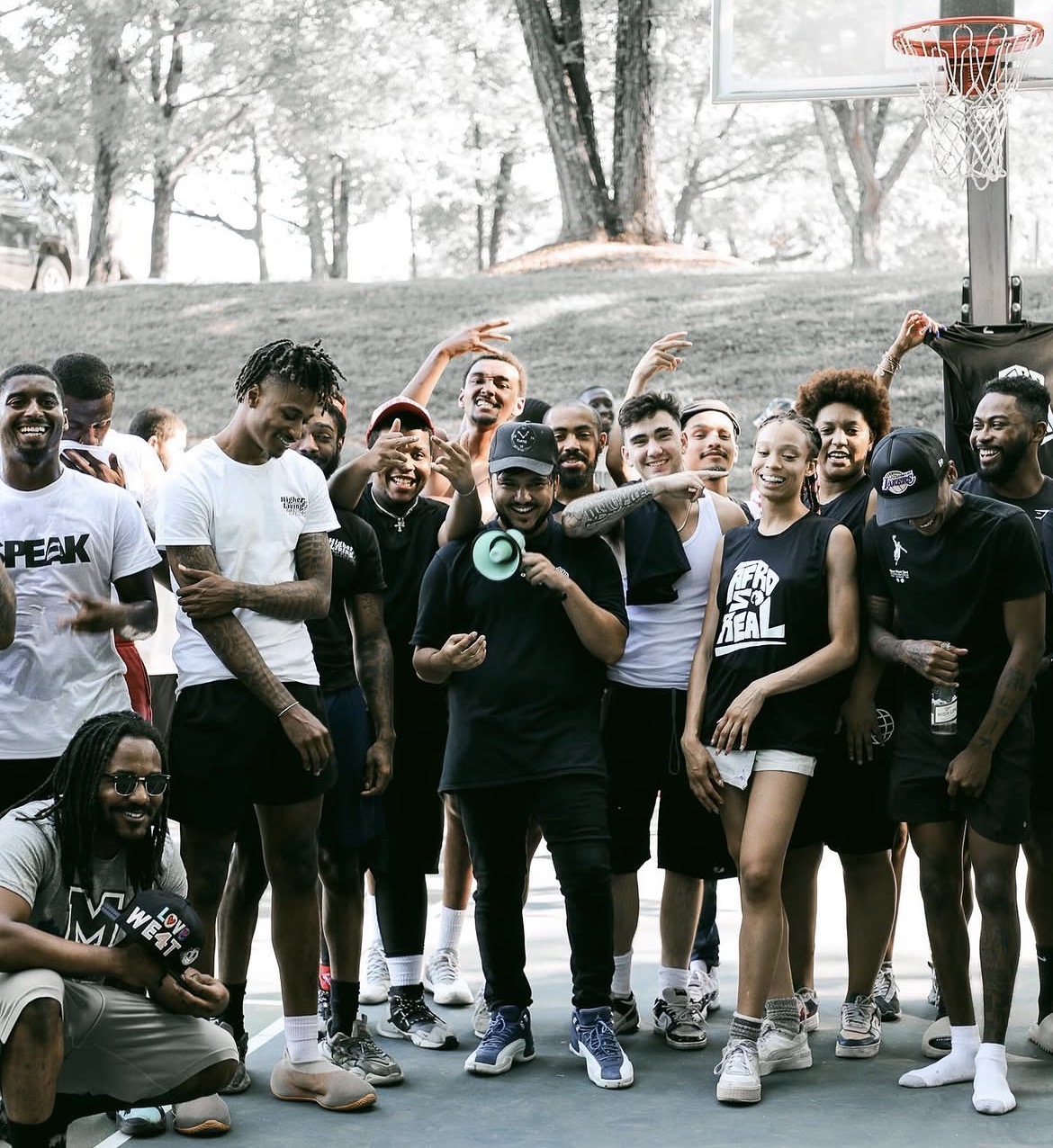 Group of young people on an outdoor basketball court, some smiling and holding drinks, with trees in the background.