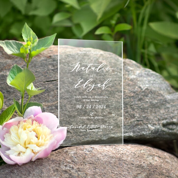 Clear acrylic sign with elegant white script text, placed on rocks with green plants and a pink flower nearby.