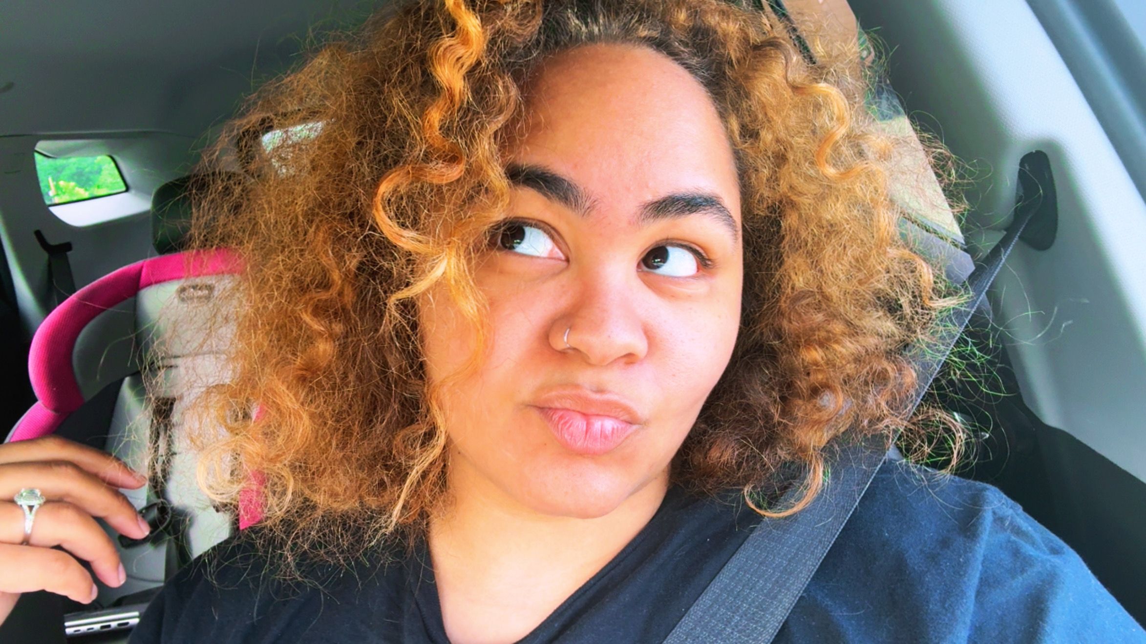 Young woman with curly hair taking a selfie inside a vehicle, holding a drink, smiling, with a pink item in the background.
