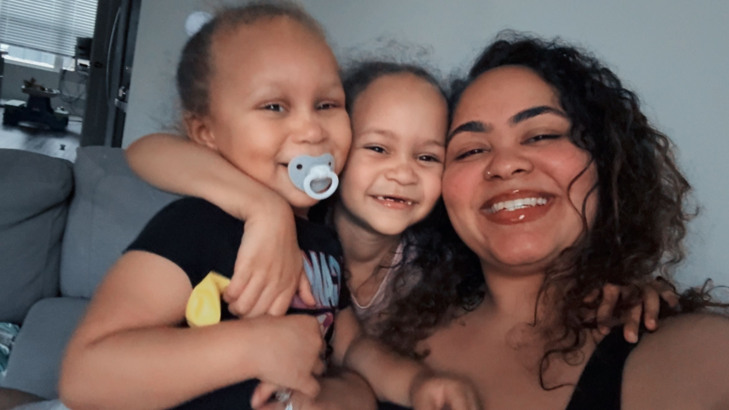 A woman with curly hair and two children, one with a pacifier, smiling together indoors.