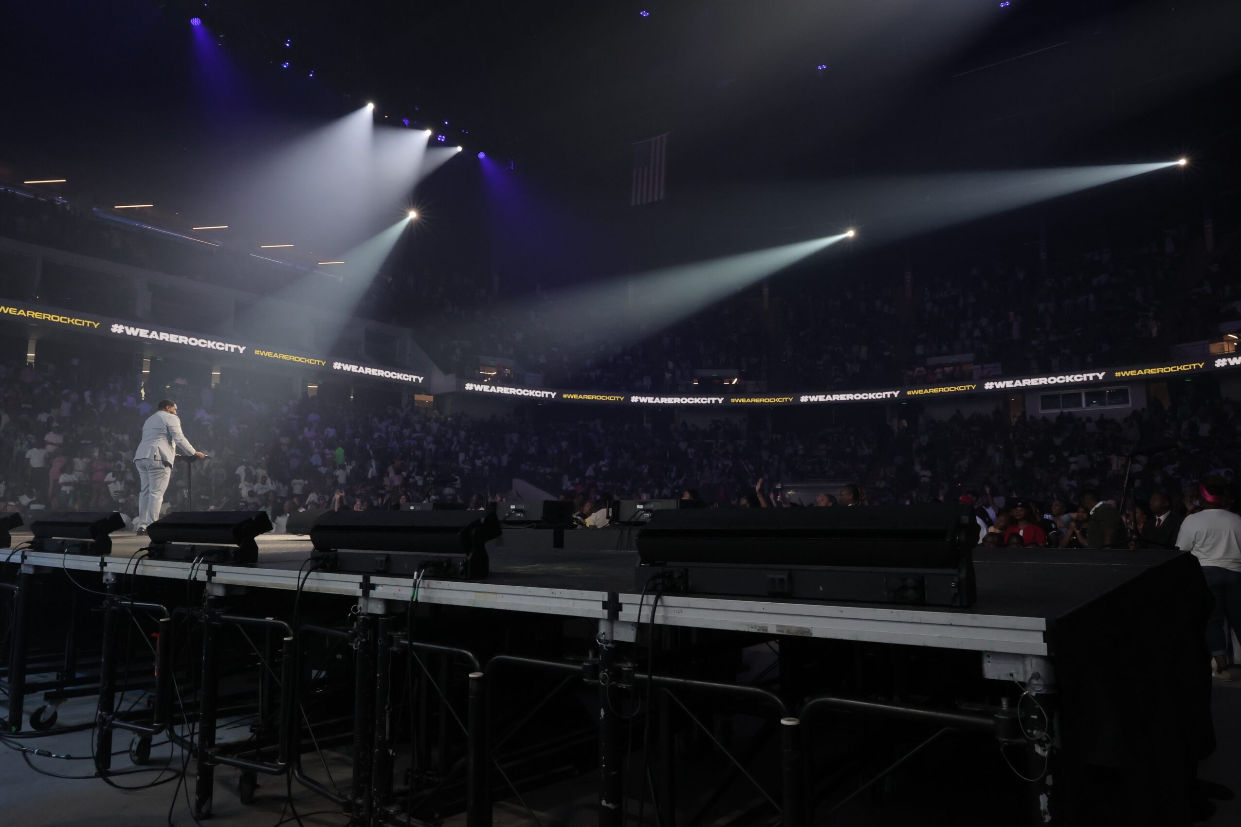 Stage with performer and audience in a large, dark arena with bright spotlights, empty chairs in foreground.