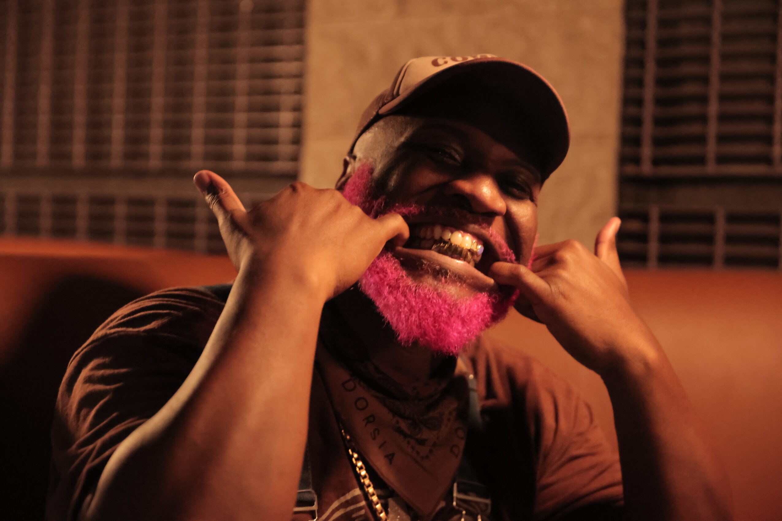 Person with pink beard smiling and making a phone gesture with hands, wearing a cap and chain, indoors at night.