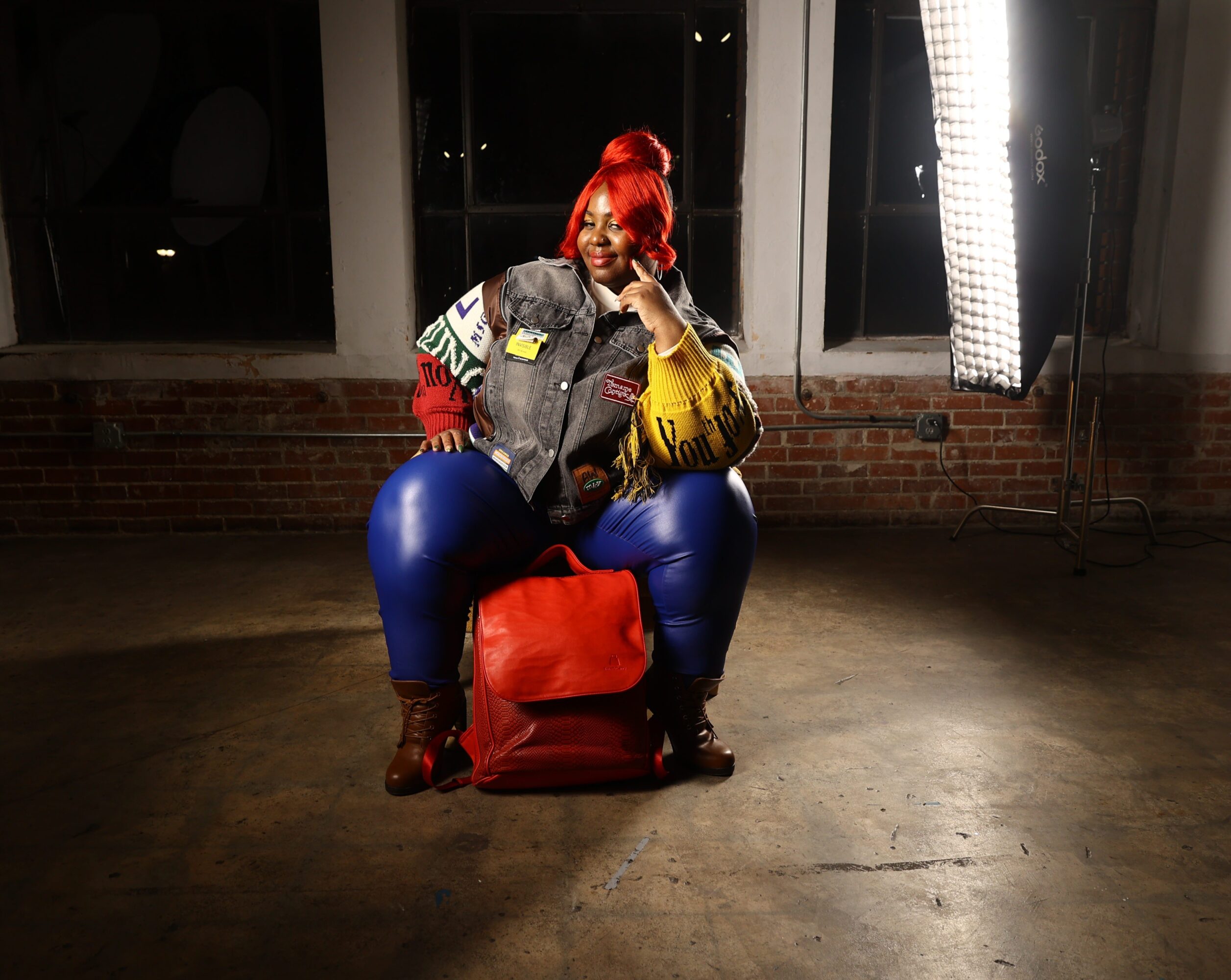 Person with red hair sitting on a red bag in a room with brick wall and large windows, illuminated by studio light.