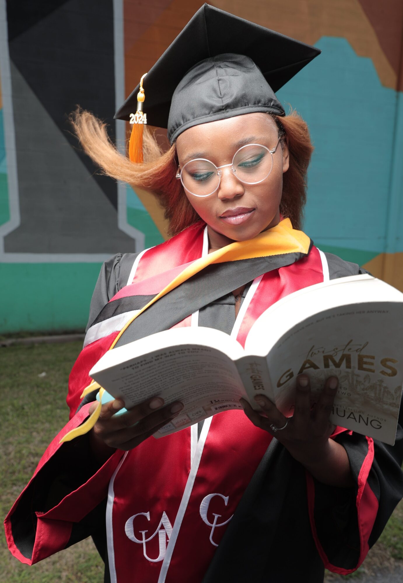 Graduate in cap and gown reading a book outdoors, with colorful mural background.