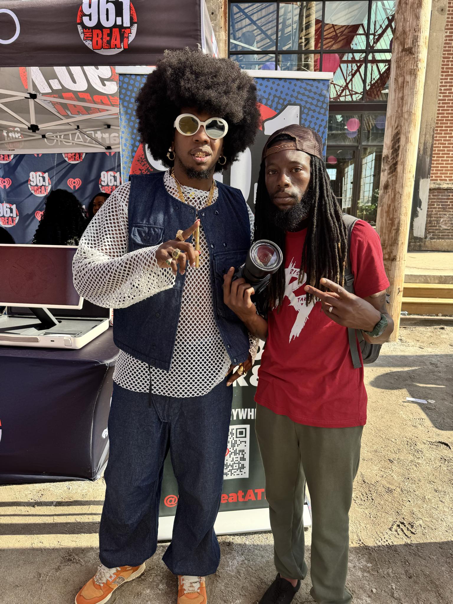 Two men standing outdoors, one with large afro hairstyle, sunglasses, and patterned clothing, the other with long dreadlocks and red shirt, holding a camera.