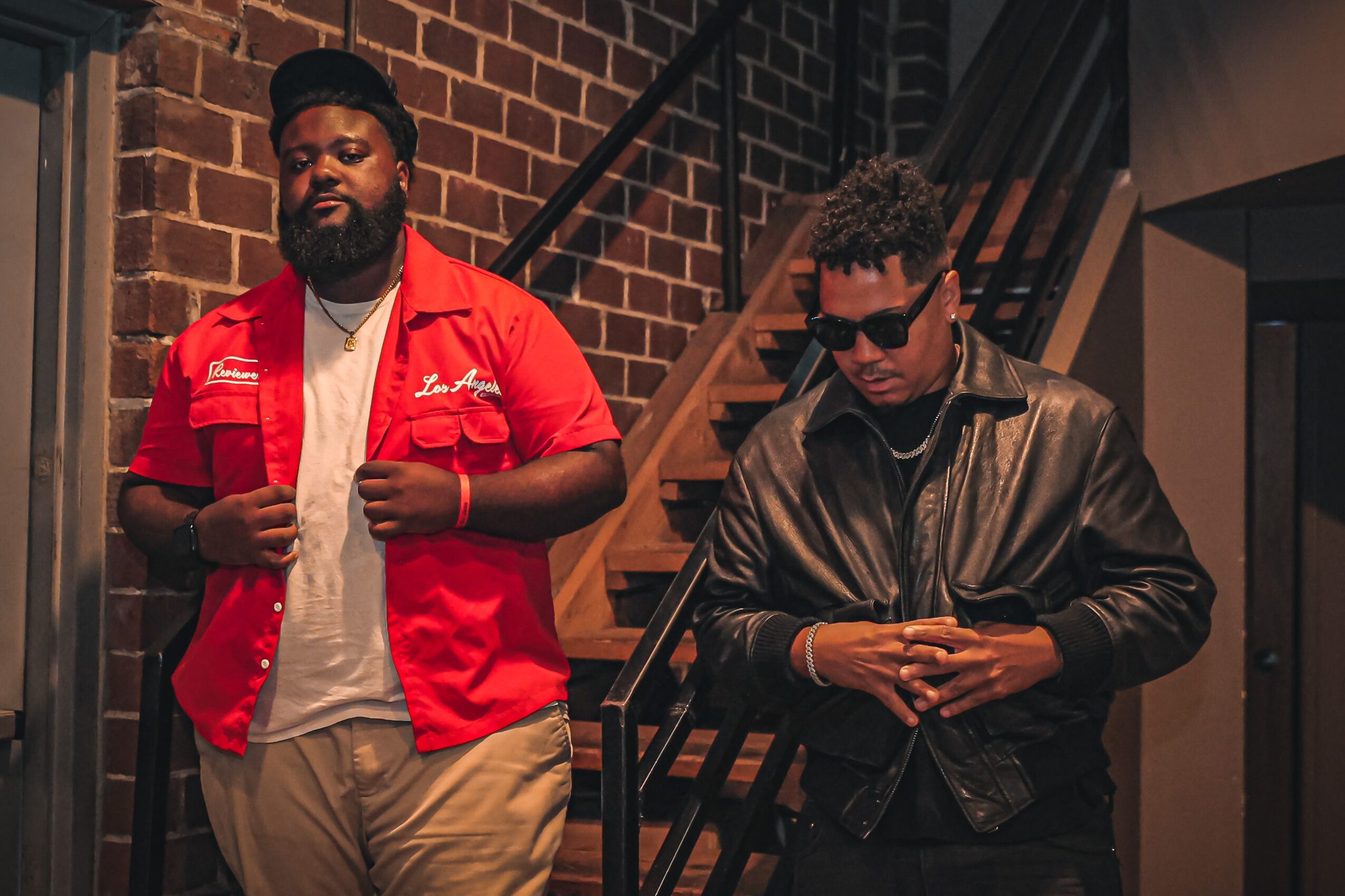 Two men standing indoors, one in a red shirt and the other in a black leather jacket, near a staircase with brick wall background.