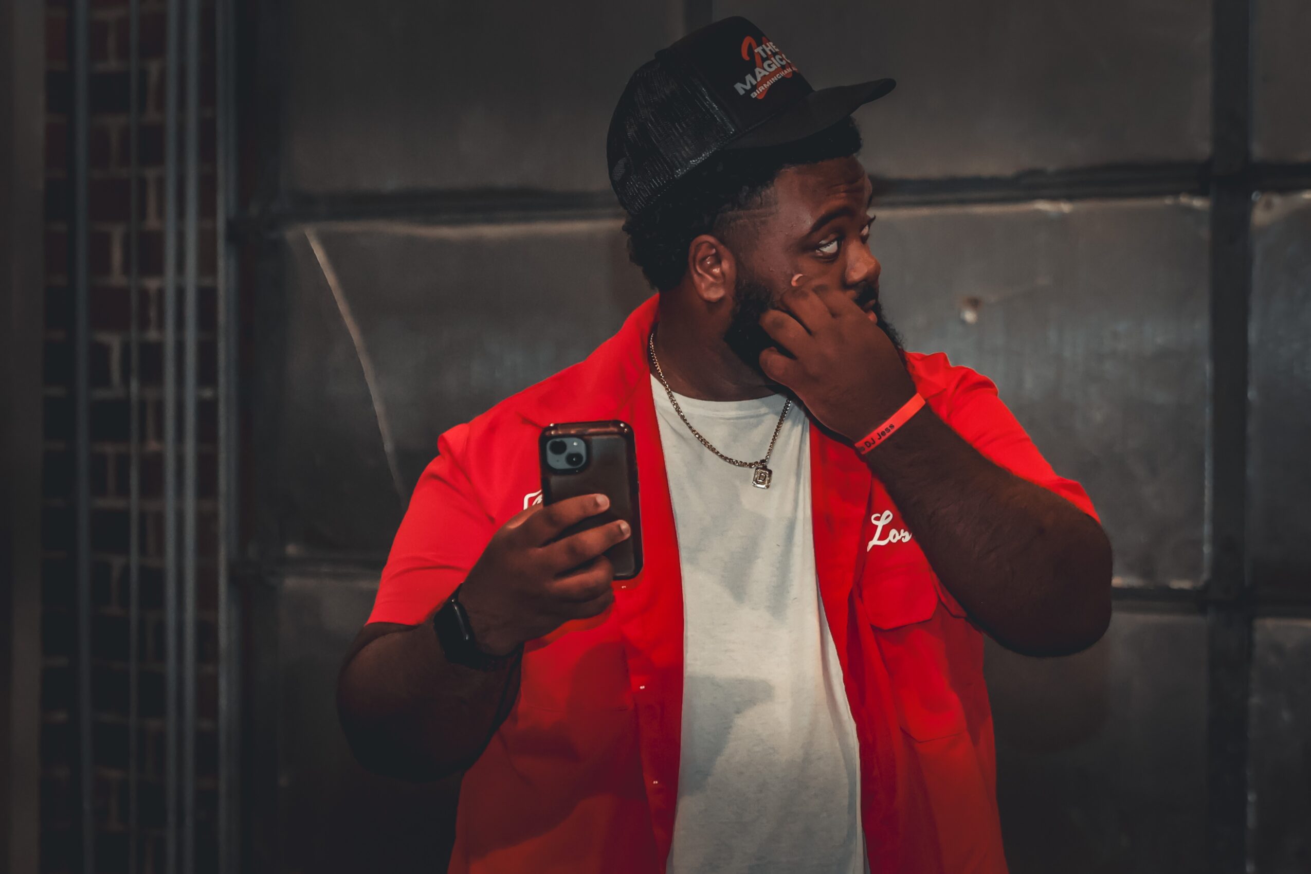 Young man with dark skin wearing a red jacket, white shirt, and black cap, holding a phone and touching his face.