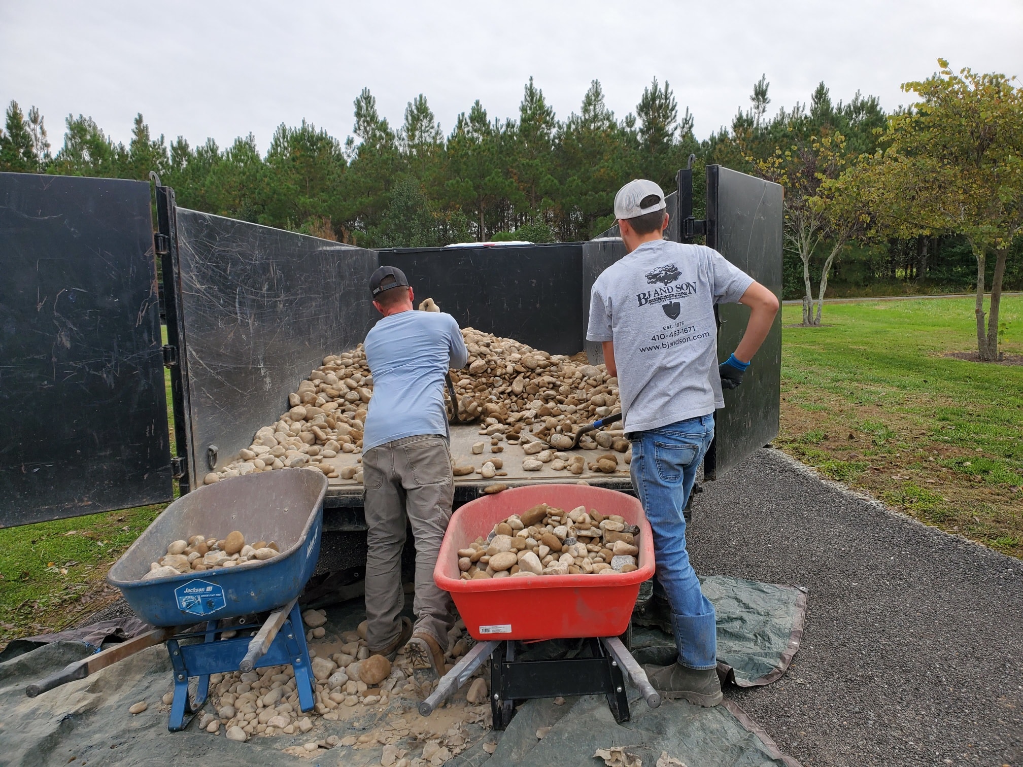 Two men are working with rocks and dirt in a large black container, with wheelbarrows nearby, on a gravel path in a park.