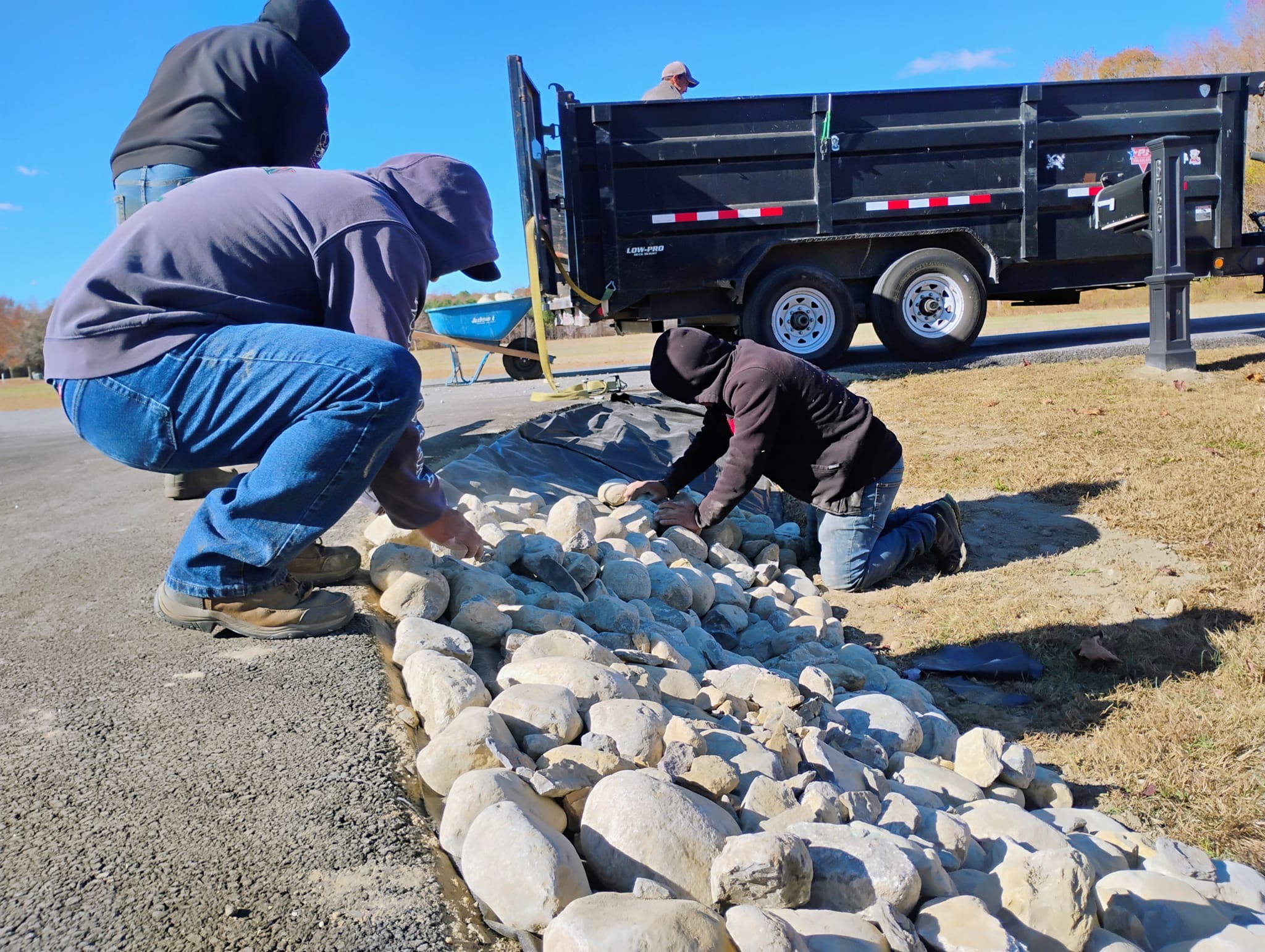 People placing rocks along a pathway with a trailer and grassy area in the background.