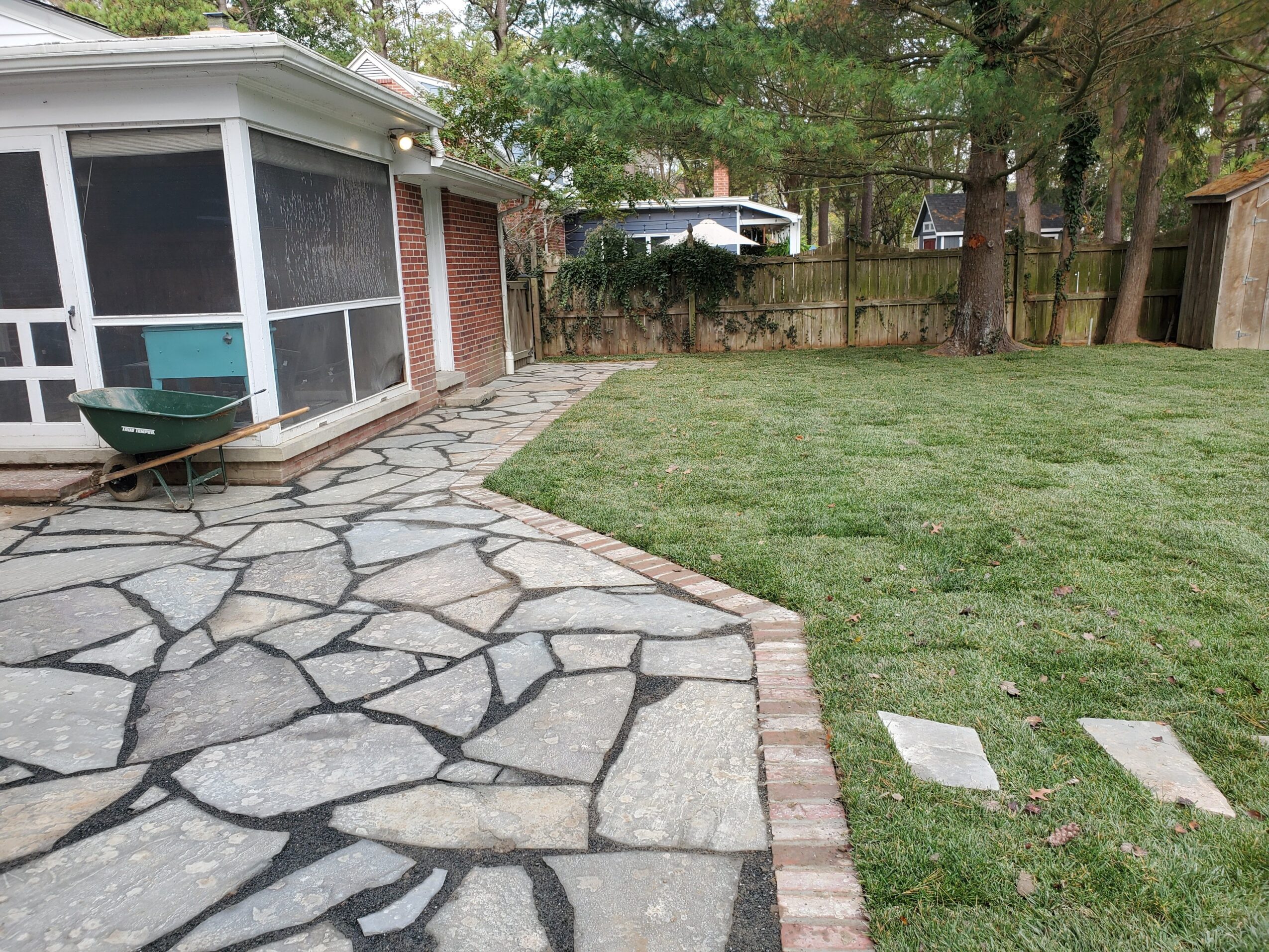 Backyard with stone patio, grassy area, screened porch, and trees in the background.