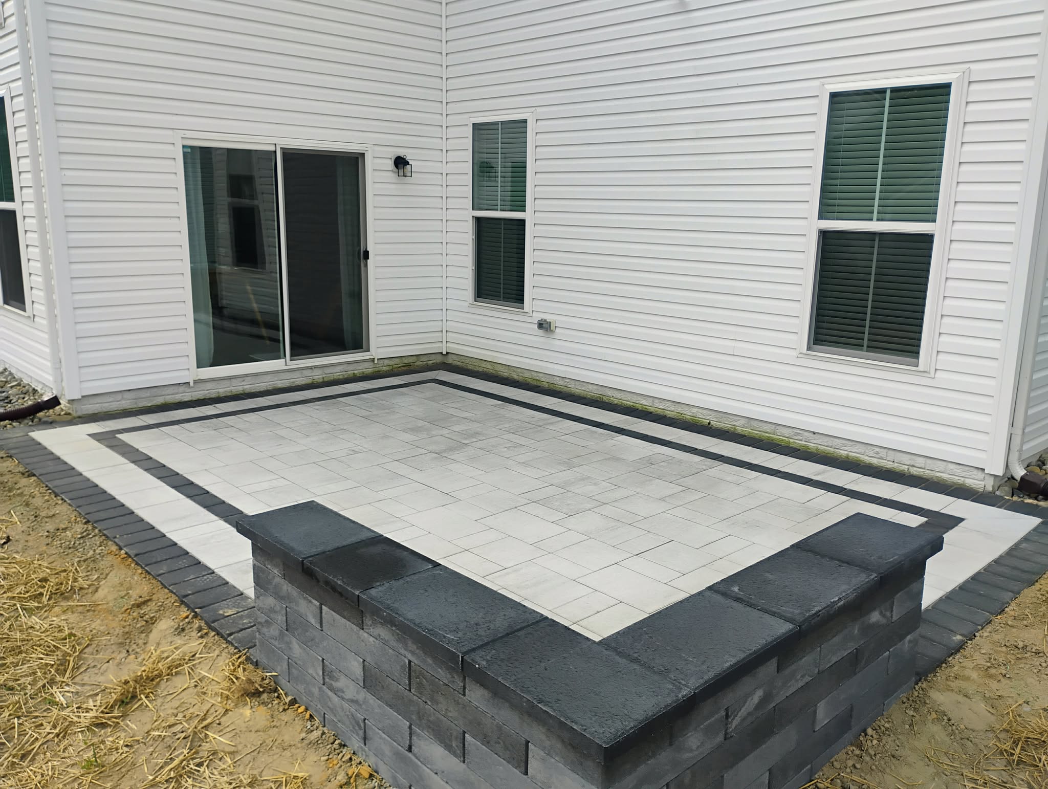 Backyard patio with gray pavers, surrounded by a black brick border, adjacent to a white house with windows.