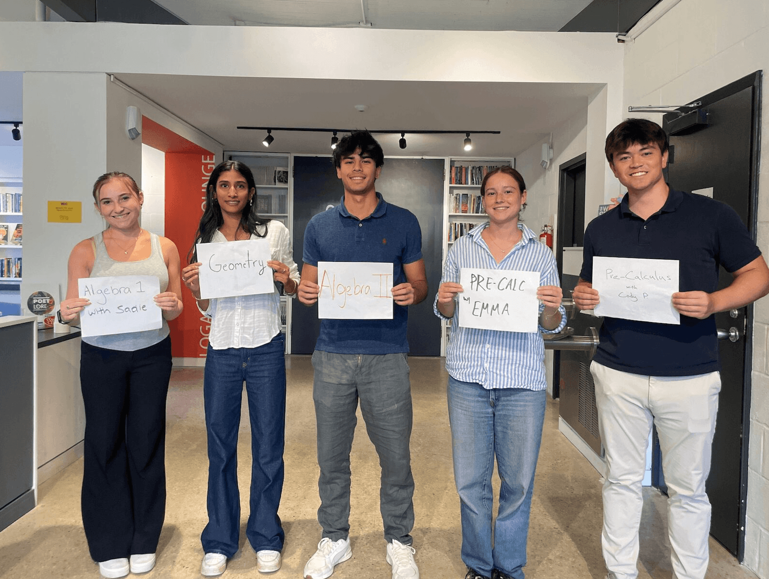 Five people standing indoors holding signs with handwritten words, smiling, in a modern room with white walls and a large screen.