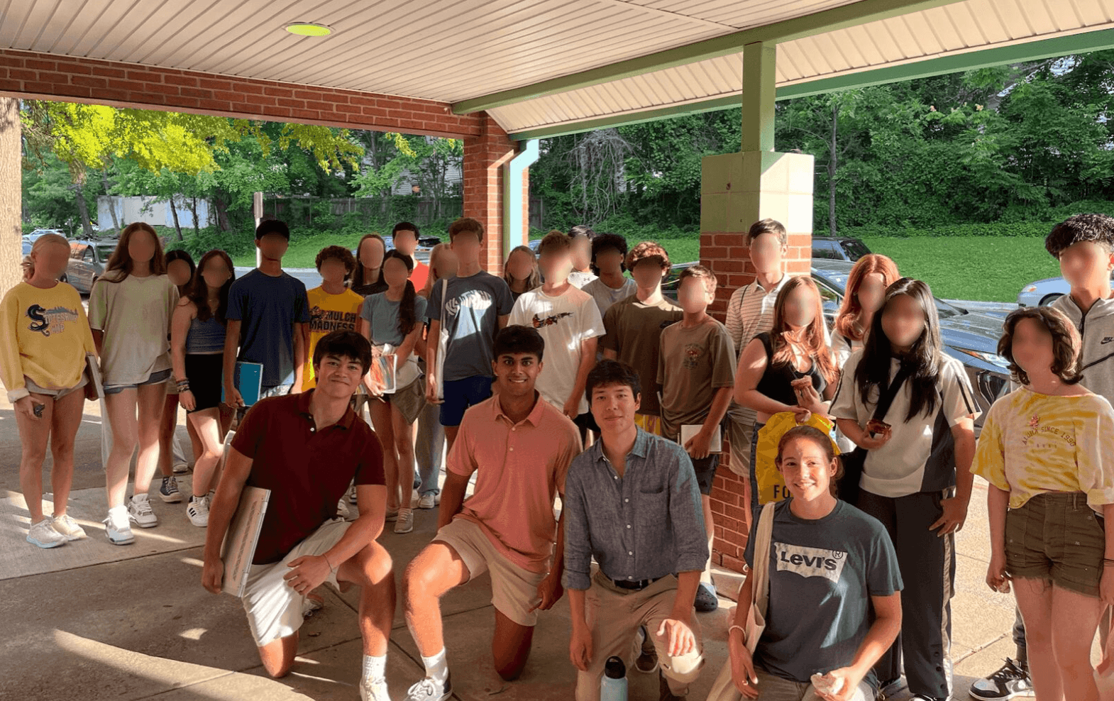Group of students under a covered outdoor area, some standing and some kneeling, with trees and a field in the background.