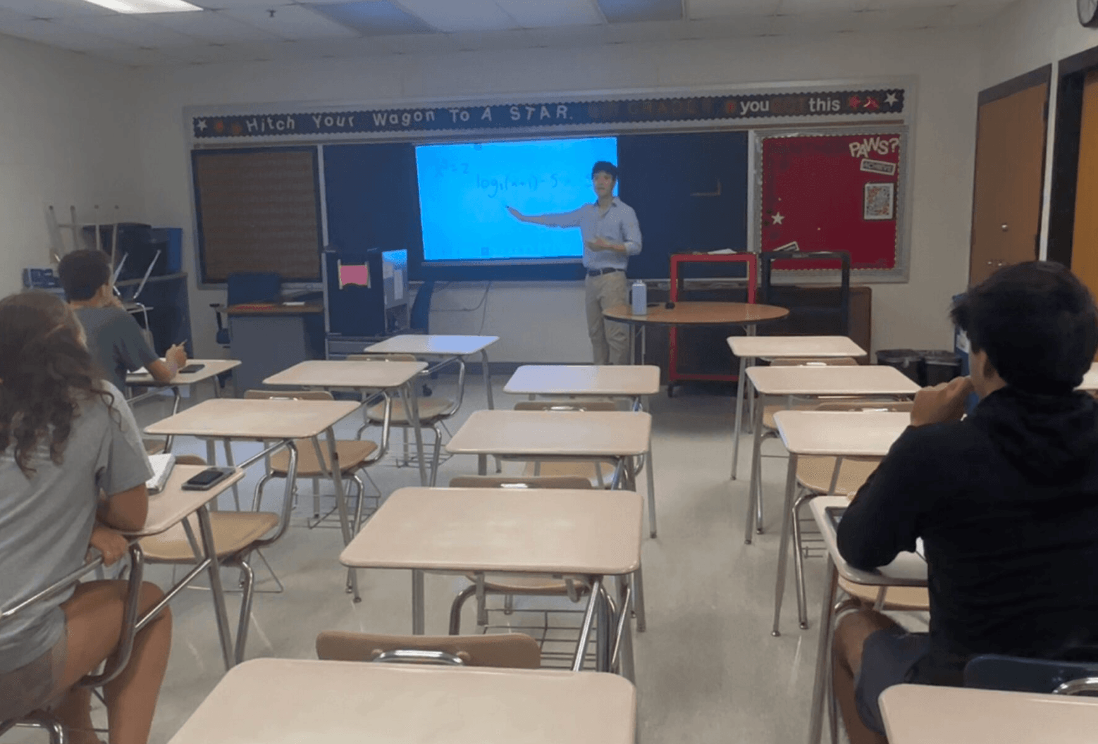 Teacher standing in front of a classroom with students seated at desks, digital screen behind teacher, classroom setting.
