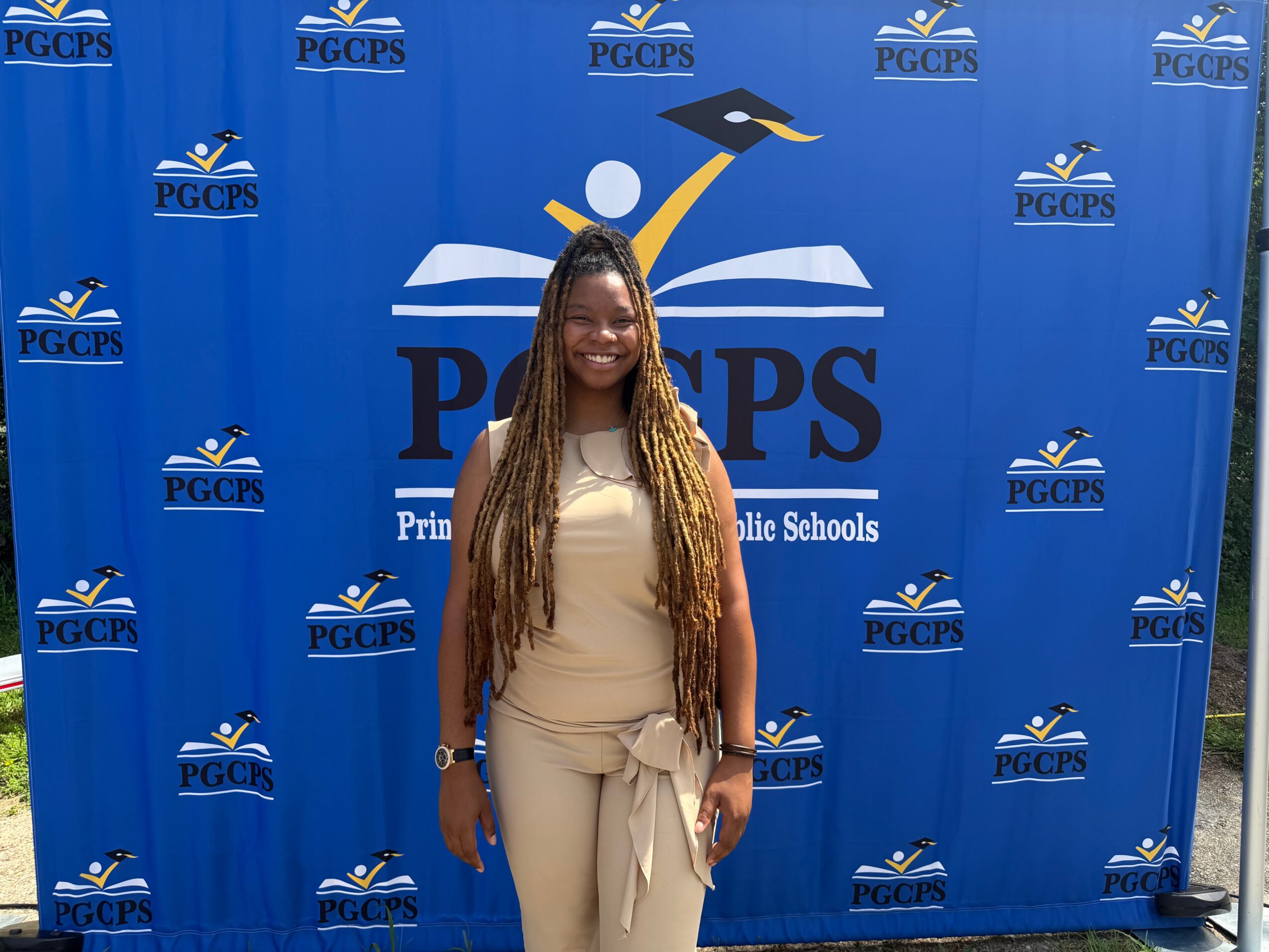 Smiling woman with long braids standing in front of a blue PGCPS backdrop at a school event.