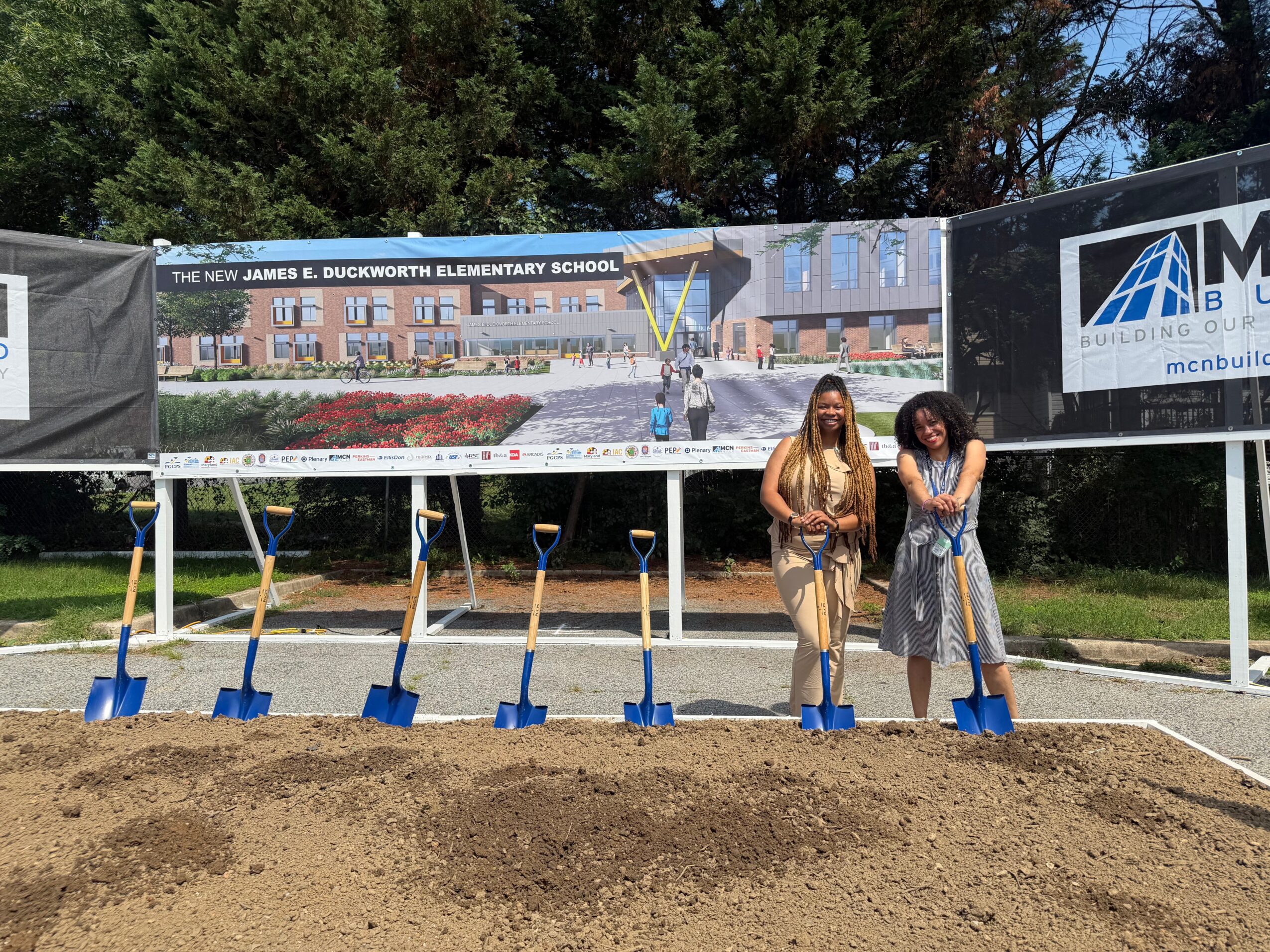 Two women stand in front of a school construction site with shovels, dirt, and a large banner, outdoors on a sunny day.