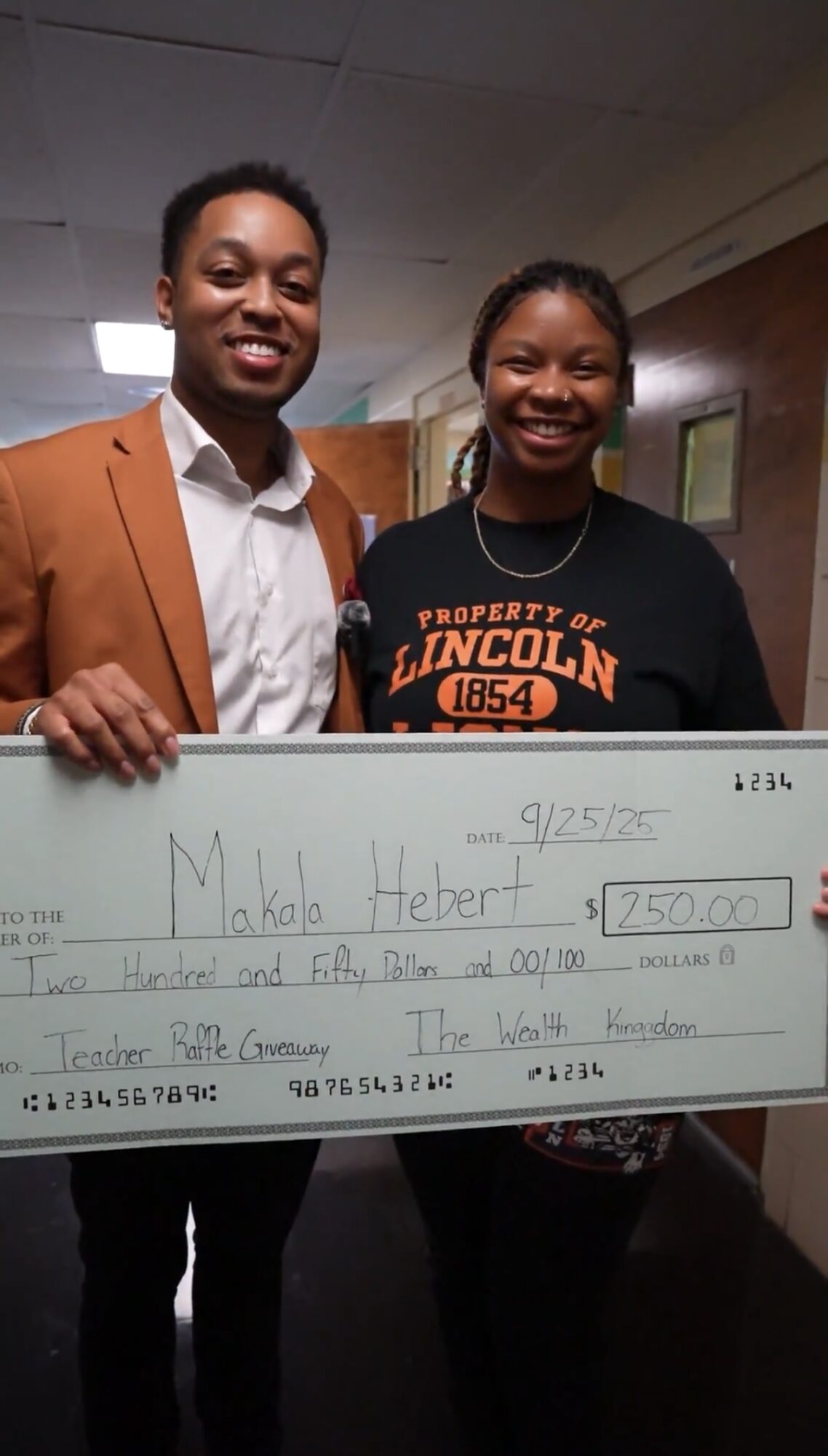 Two smiling people holding a large check, one wearing a brown blazer and the other a black shirt with orange text.