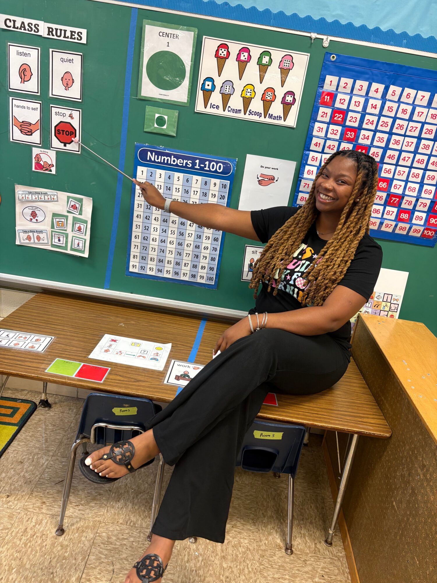 Woman standing next to a classroom bulletin board with educational posters and charts, pointing at a chart with a pointer.