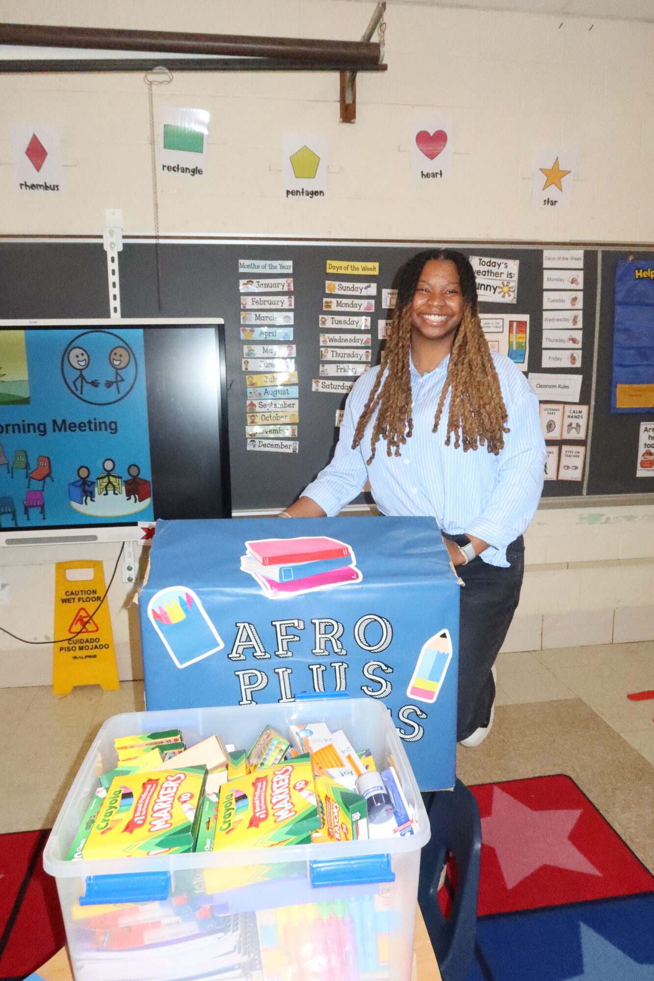Woman standing behind a table with school supplies in a classroom, shelves and decorations in the background.