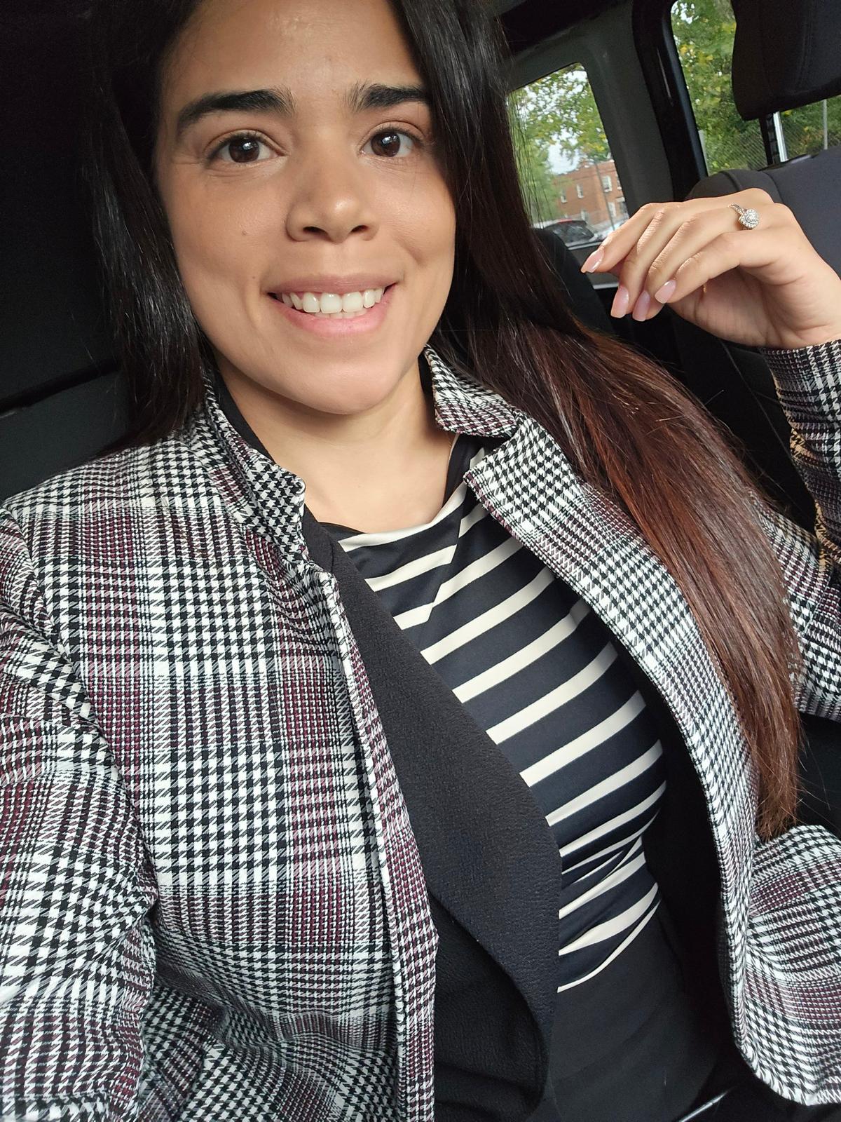 Young woman with long dark hair smiling, wearing a plaid jacket and striped shirt, sitting in a car.