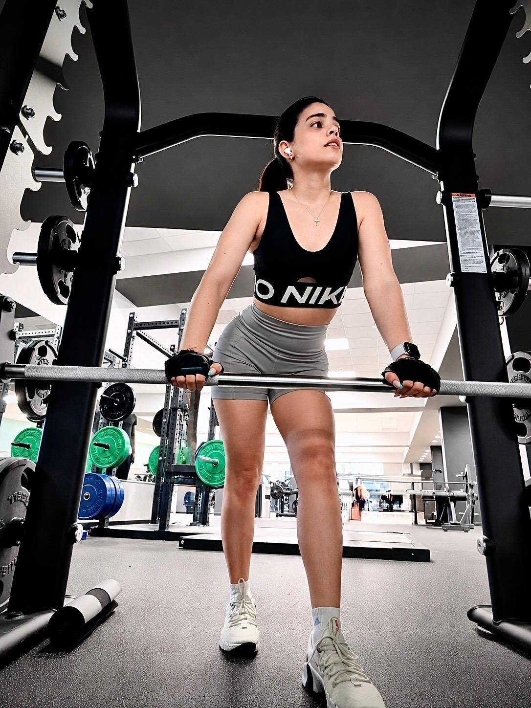 Young woman in workout clothes lifting a barbell in a gym, looking upward, with weight racks in background.