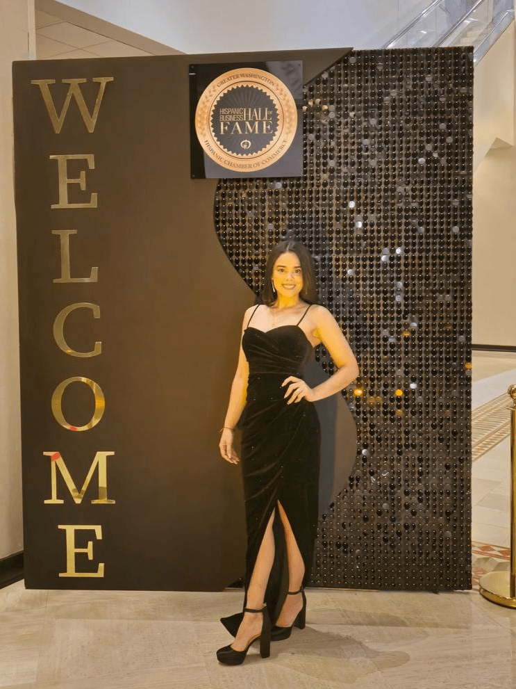 Woman in black dress with slit, standing in front of a welcome sign and decorative backdrop, posing with hand on hip.